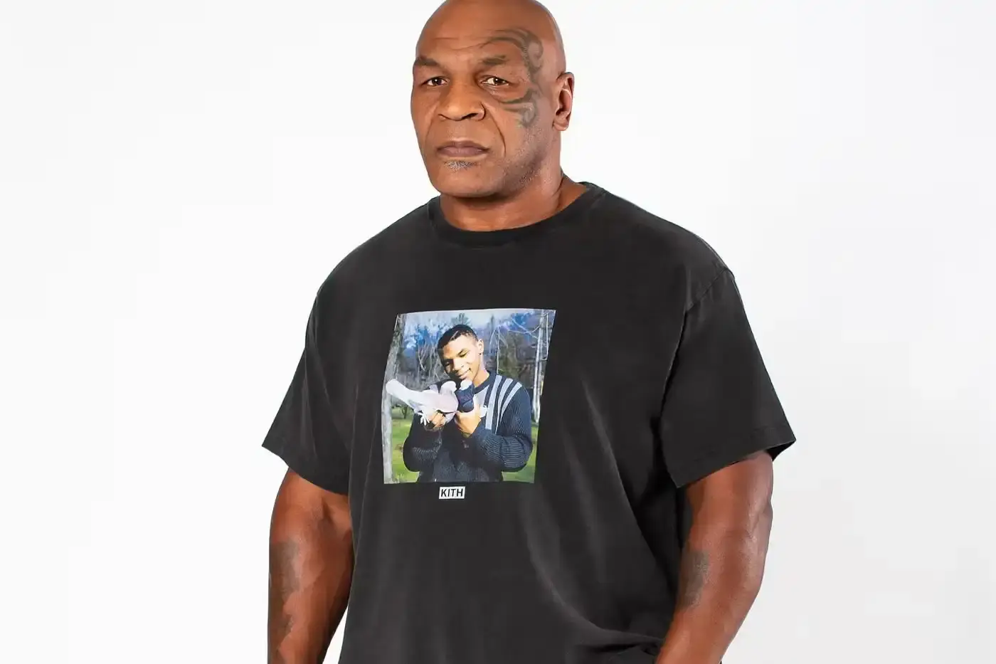 Kith The image shows Mike Tyson, the famous boxer, gently pressing two pigeons together as if they are kissing. This is a recreation of the "Now Kith" meme, which references Tyson's distinctive lisp. Tyson is wearing one of the collaborative t-shirts from his Kith collection, which features the meme image in a vintage-inspired design.