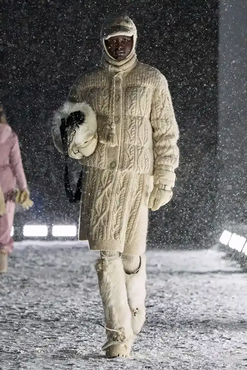 Moncler Moncler's Fall/Winter 2025 Grenoble collection showcase held at Courchevel Altiport in the French Alps. It details how the runway show transformed Europe's highest airport into a fashion venue at 2,008 meters above sea level. The article outlines the collection's 140 looks that blend urban style with mountain functionality, featuring innovative materials like denim and canvas alongside traditional winter textiles. It mentions the craftsmanship details including embroidery and intarsia patterns on ski suits. The article also notes celebrity attendees and a special snowboard collaboration with Shaun White's WHITESPACE brand. The content is organized in paragraphs describing the event setting, collection details, and notable highlights (image 18 of 22)