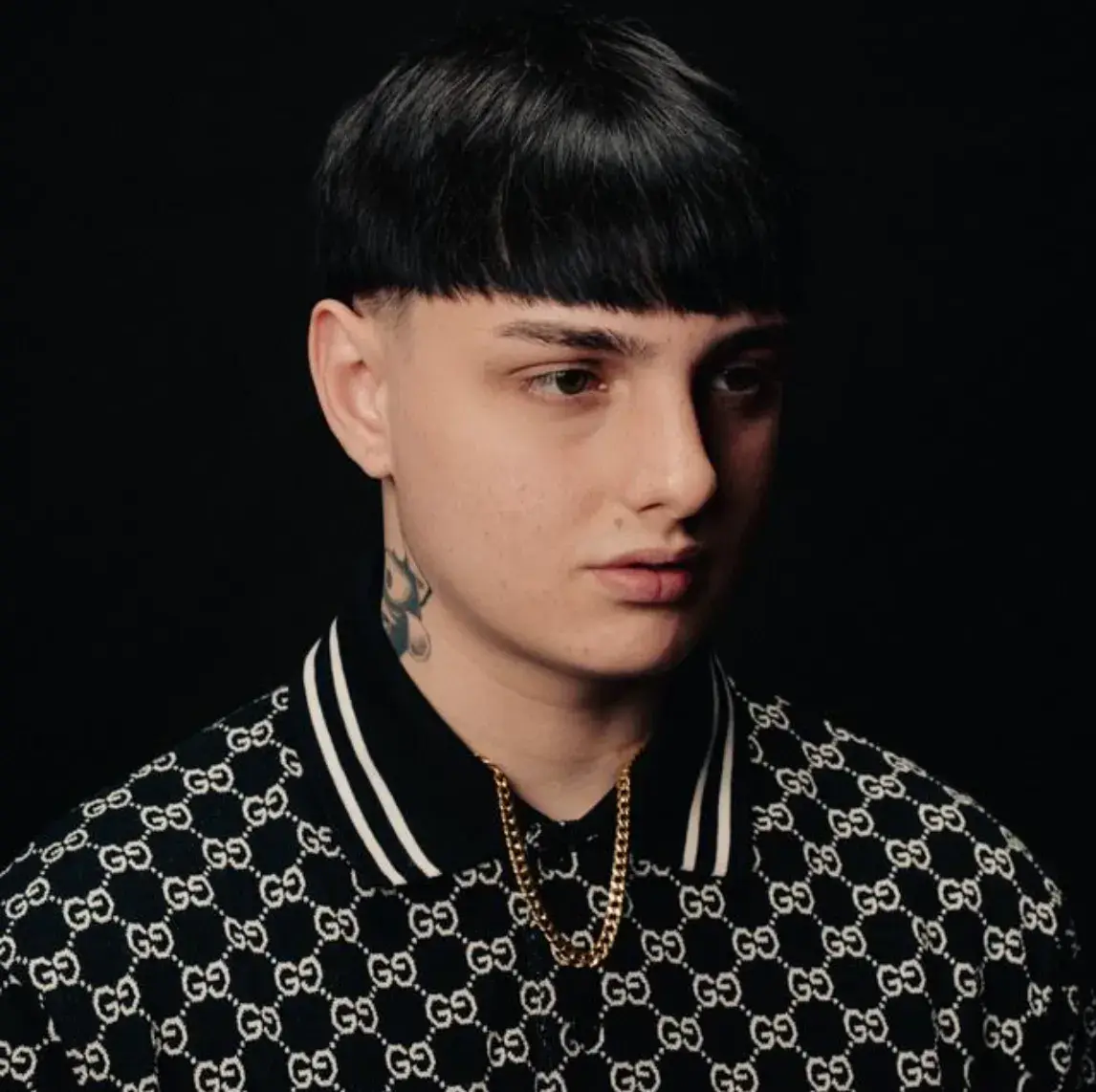 Italian rapper Dagood YL in a black monogram shirt and gold chain against a dark background.