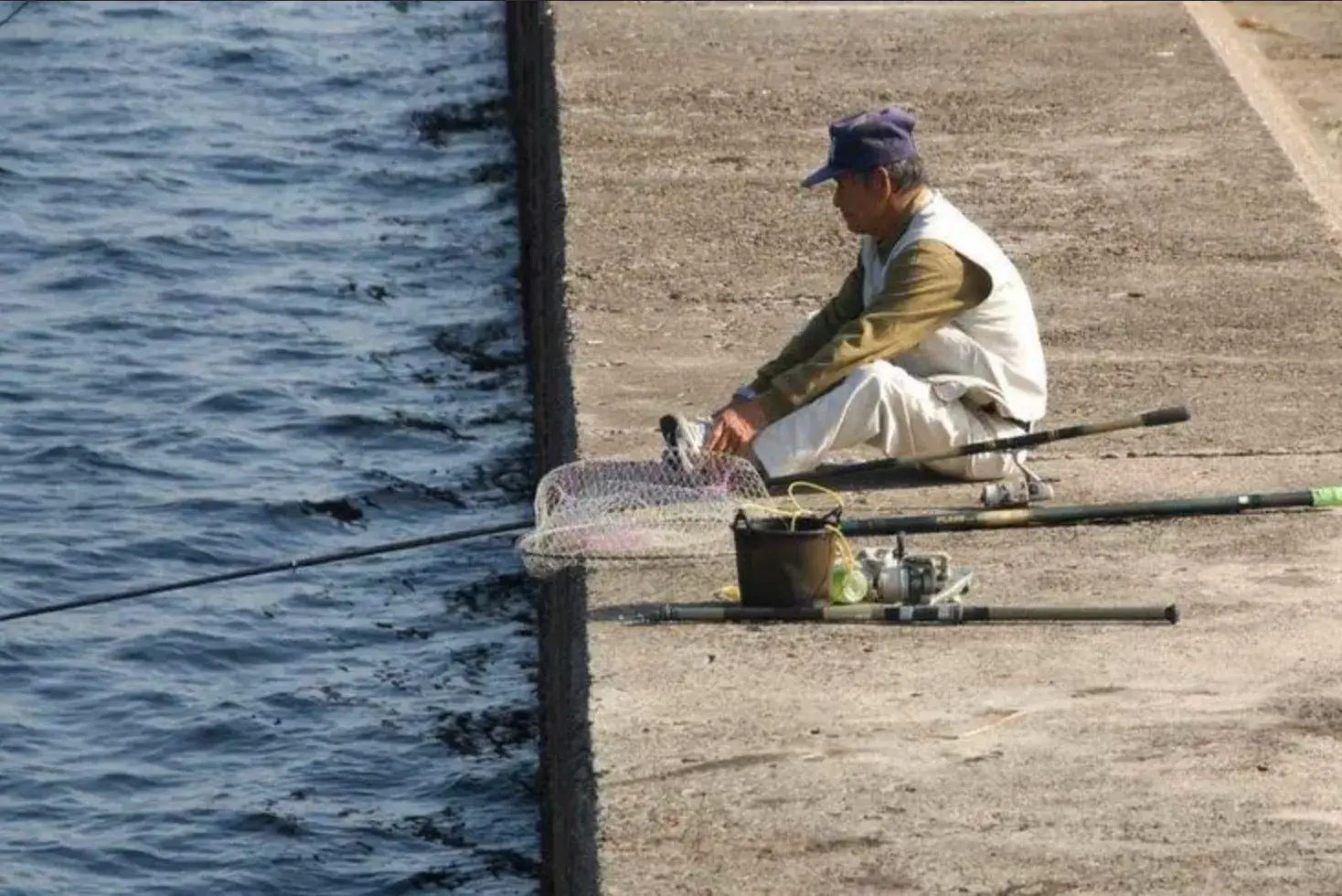 Elderly fisherman sitting on a concrete pier beside the sea, with fishing rods, a landing net and a bucket