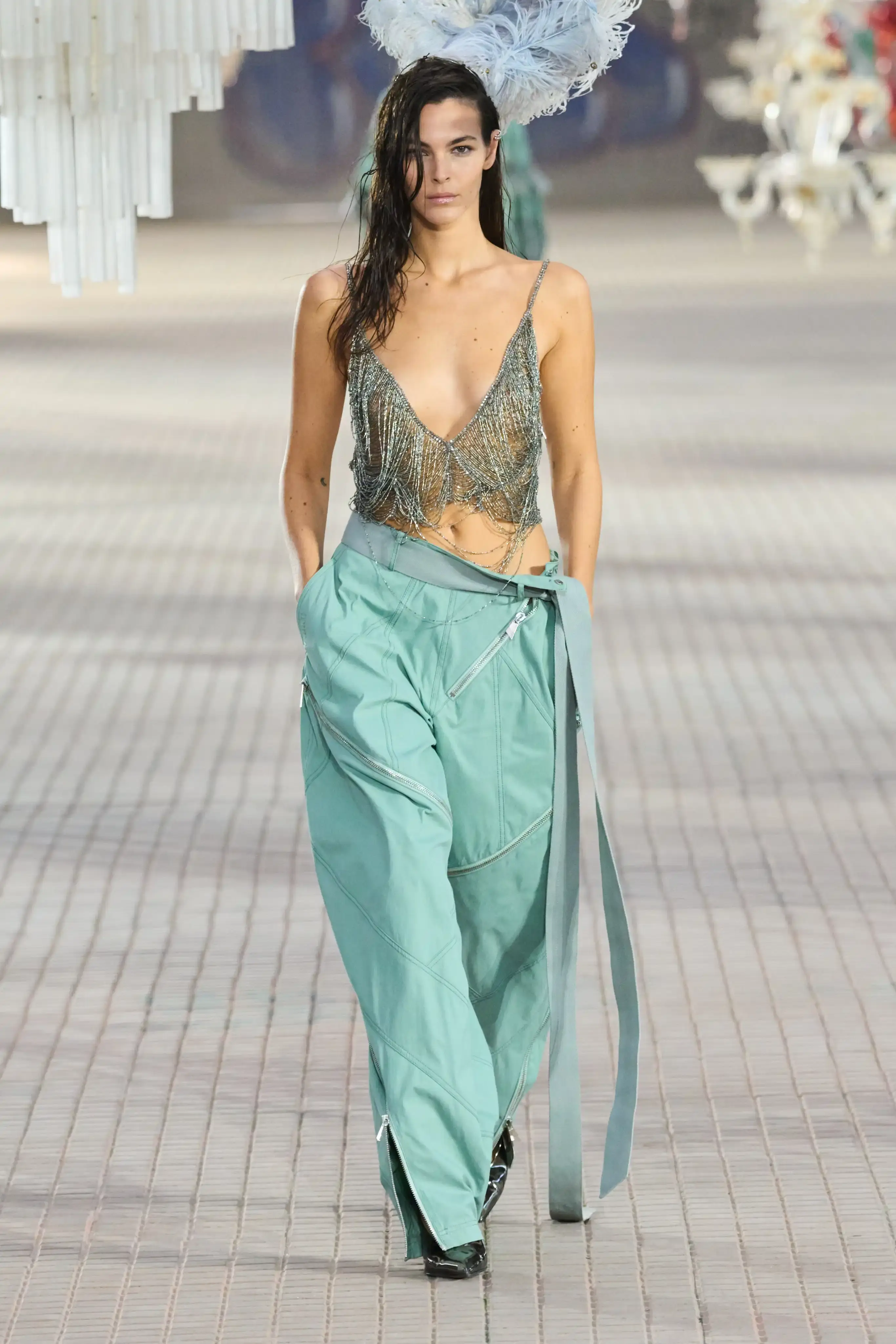 The Attico's Spring/Summer 2025 runway show at Milan Fashion Week features a surprising collaboration with Nike. Models walk on an industrial set adorned with colorful glass chandeliers, showcasing a collection that blends high fashion with sportswear elements. The runway presentation, titled "The Sound of Breaking Glass," explores themes of fragility and strength through contrasting designs. Sheer fabrics, form-fitting silhouettes, and embroideries are juxtaposed with nylon materials, bomber jackets, and cargo pants. The color palette is predominantly dusty and faded. Select Nike athletic wear pieces are integrated into the collection, symbolizing resilience and personal growth. Designers Gilda Ambrosio and Giorgia Tordini aim to empower women with this innovative fusion of luxury fashion and athletic functionality, challenging traditional runway norms (image 2 of 6)