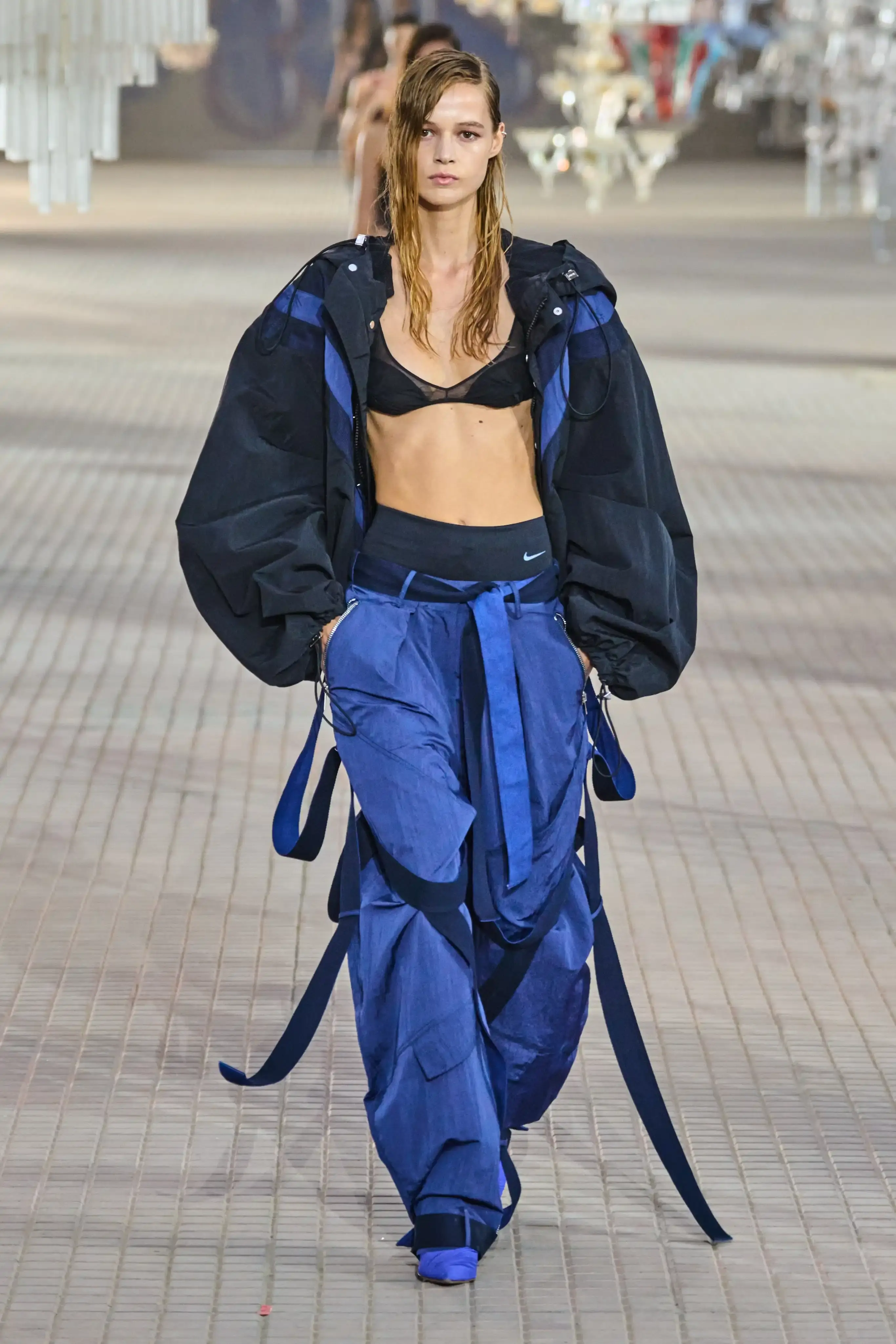 The Attico's Spring/Summer 2025 runway show at Milan Fashion Week features a surprising collaboration with Nike. Models walk on an industrial set adorned with colorful glass chandeliers, showcasing a collection that blends high fashion with sportswear elements. The runway presentation, titled "The Sound of Breaking Glass," explores themes of fragility and strength through contrasting designs. Sheer fabrics, form-fitting silhouettes, and embroideries are juxtaposed with nylon materials, bomber jackets, and cargo pants. The color palette is predominantly dusty and faded. Select Nike athletic wear pieces are integrated into the collection, symbolizing resilience and personal growth. Designers Gilda Ambrosio and Giorgia Tordini aim to empower women with this innovative fusion of luxury fashion and athletic functionality, challenging traditional runway norms (image 4 of 6)