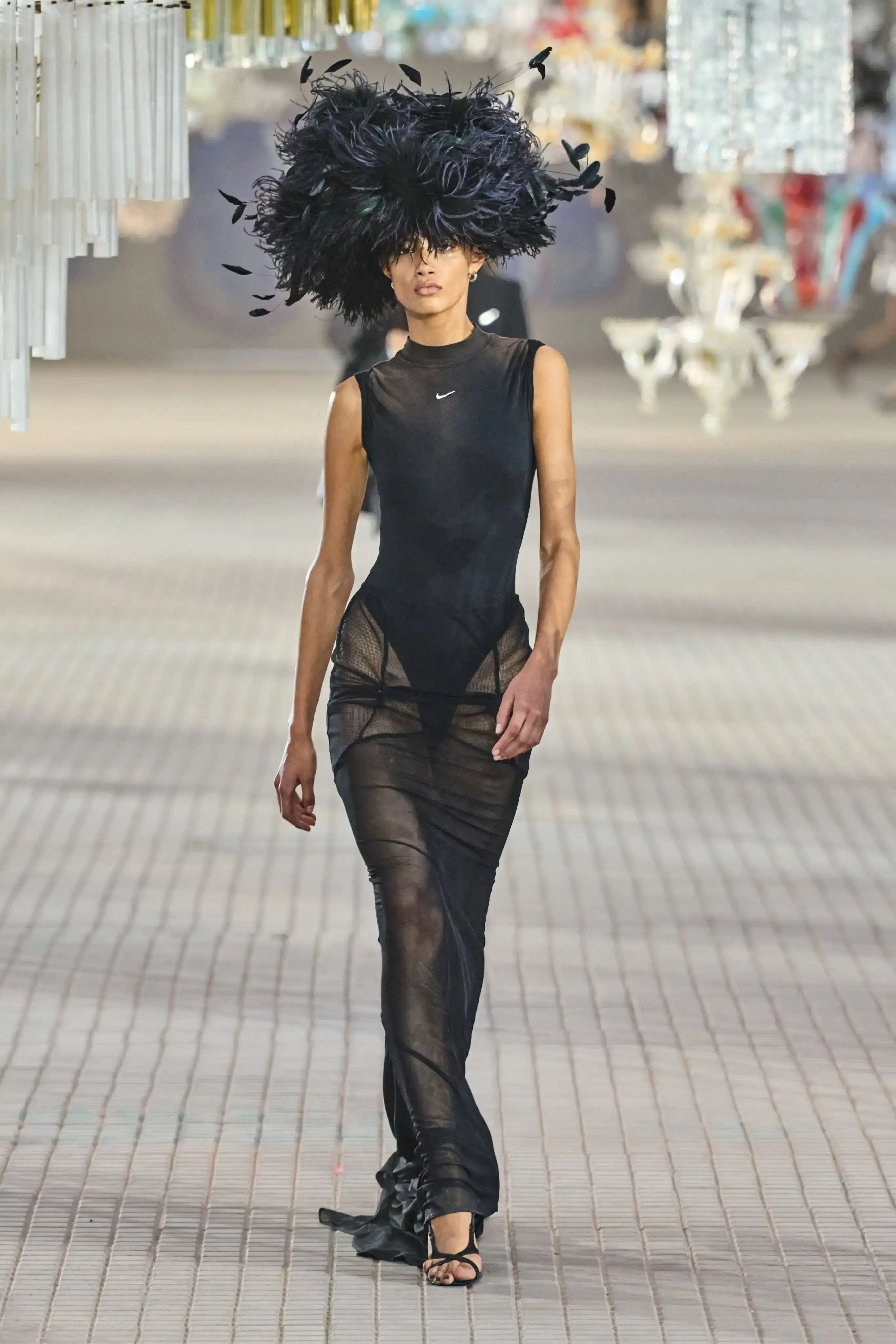 The Attico's Spring/Summer 2025 runway show at Milan Fashion Week features a surprising collaboration with Nike. Models walk on an industrial set adorned with colorful glass chandeliers, showcasing a collection that blends high fashion with sportswear elements. The runway presentation, titled "The Sound of Breaking Glass," explores themes of fragility and strength through contrasting designs. Sheer fabrics, form-fitting silhouettes, and embroideries are juxtaposed with nylon materials, bomber jackets, and cargo pants. The color palette is predominantly dusty and faded. Select Nike athletic wear pieces are integrated into the collection, symbolizing resilience and personal growth. Designers Gilda Ambrosio and Giorgia Tordini aim to empower women with this innovative fusion of luxury fashion and athletic functionality, challenging traditional runway norms (image 5 of 6)
