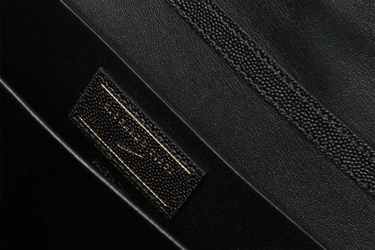 Nike x Riccardo Tisci The Mercer Bag by Nike x Riccardo Tisci, shown in black pebbled leather with gold hardware. The minimalist design features a front flap with debossed Nike logo and R.T. metal signature at the corner. The bag includes an adjustable shoulder strap. (image 5 of 6)