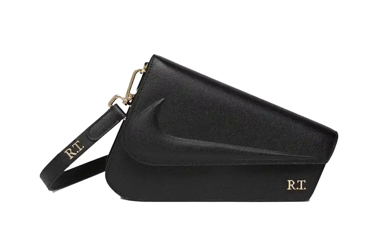 Nike x Riccardo Tisci The Mercer Bag by Nike x Riccardo Tisci, shown in black pebbled leather with gold hardware. The minimalist design features a front flap with debossed Nike logo and R.T. metal signature at the corner. The bag includes an adjustable shoulder strap. (image 2 of 6)