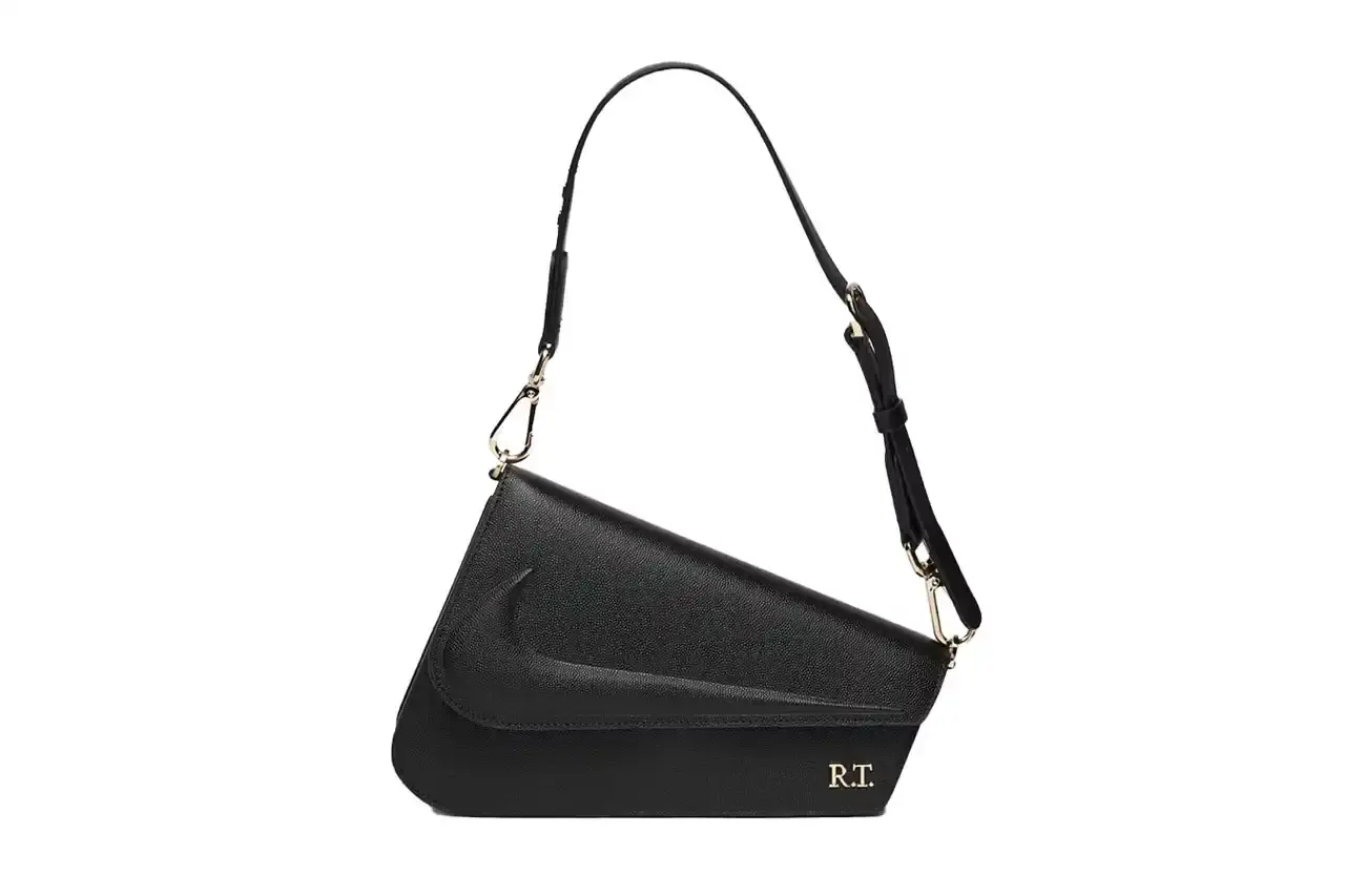 Nike x Riccardo Tisci The Mercer Bag by Nike x Riccardo Tisci, shown in black pebbled leather with gold hardware. The minimalist design features a front flap with debossed Nike logo and R.T. metal signature at the corner. The bag includes an adjustable shoulder strap.
