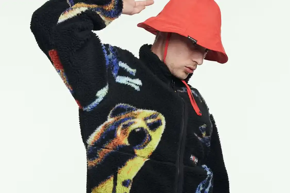 Article about Palace skateboard brand showing preview image of upcoming Autumn 2025 clothing collection. Main image shows person wearing black zip-up sherpa jacket decorated with blue, yellow, and orange frog graphics, brown camouflage pants, and orange bucket hat. Article discusses Palace's recent releases and provides context about the brand's seasonal collection strategy.
