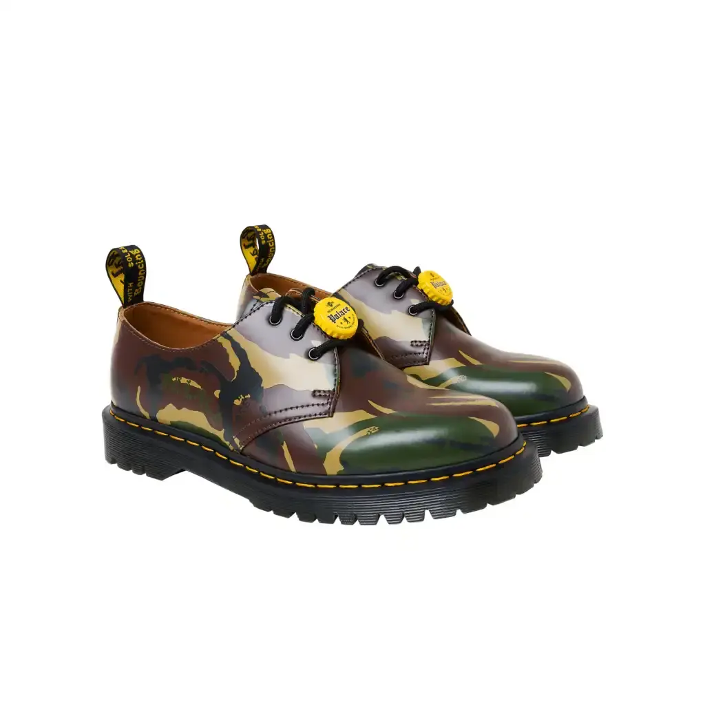 PALACE TEAMS UP WITH DR. MARTENS FOR FIRST-EVER COLLAB photo of the first collaborative collection between British brands Palace and Dr. Martens. The collection features three versions of Dr. Martens' classic 1461 shoe in black, cherry red, and woodland camouflage colorways. Each pair is made from vintage leather and includes distinctive bottle cap charms inspired by 1980s trends. The shoes feature a BEN sole with traditional yellow stitching and "Palace" text on the heel loop instead of the usual AirWair logo. The collection will be available online starting April 11 at 12:00 (image 2 of 3)