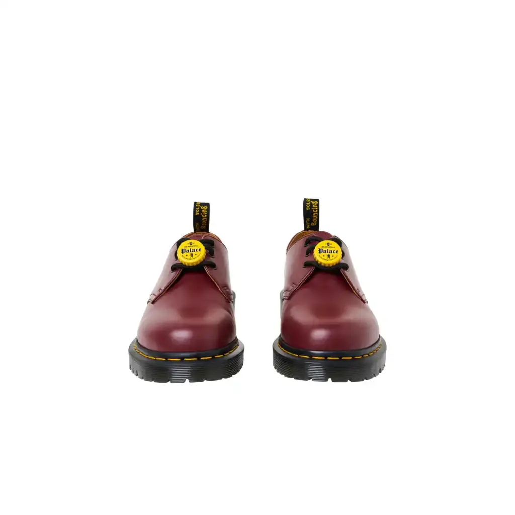 PALACE TEAMS UP WITH DR. MARTENS FOR FIRST-EVER COLLAB photo of the first collaborative collection between British brands Palace and Dr. Martens. The collection features three versions of Dr. Martens' classic 1461 shoe in black, cherry red, and woodland camouflage colorways. Each pair is made from vintage leather and includes distinctive bottle cap charms inspired by 1980s trends. The shoes feature a BEN sole with traditional yellow stitching and "Palace" text on the heel loop instead of the usual AirWair logo. The collection will be available online starting April 11 at 12:00 (image 3 of 3)