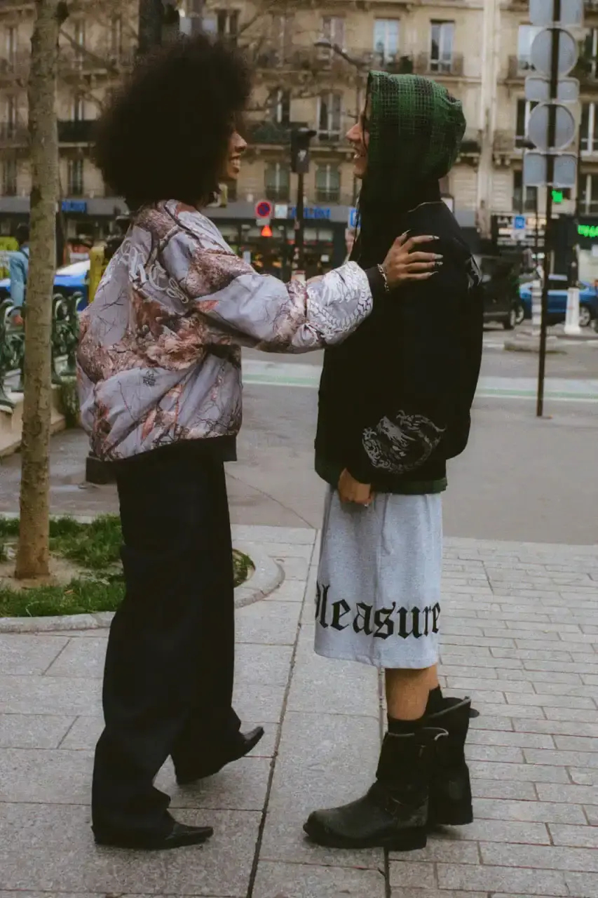 Quinn Dunziellas PLEASURES Spring/Summer 2025 lookbook photograph showcases models wearing the new collection against Parisian backdrops. Featured items include a Realtree camo jacket, leopard print blazer, and holographic-lined outerwear pieces. (image 14 of 15)