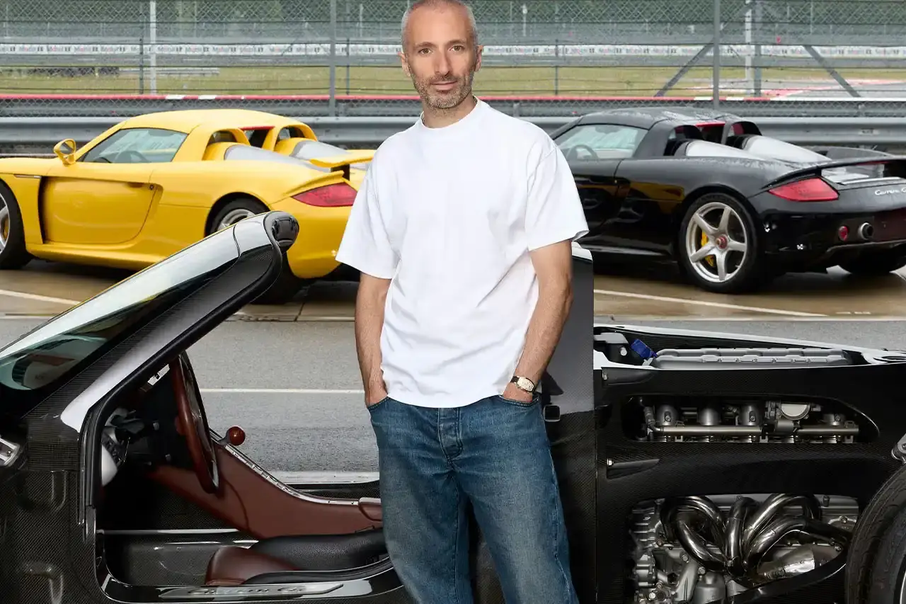 image reports on a fashion collaboration between German automaker Porsche and Paris-based designer Arthur Kar. The piece covers their upcoming September 2025 collection celebrating the 25th anniversary of the Carrera GT supercar. It includes background information on their previous partnerships, details about the Carrera GT's specifications and production history, and quotes from both Kar and Porsche executives. The article is structured with a summary section followed by detailed reporting on the collaboration, the car's significance, and the companies' broader lifestyle marketing strategy.