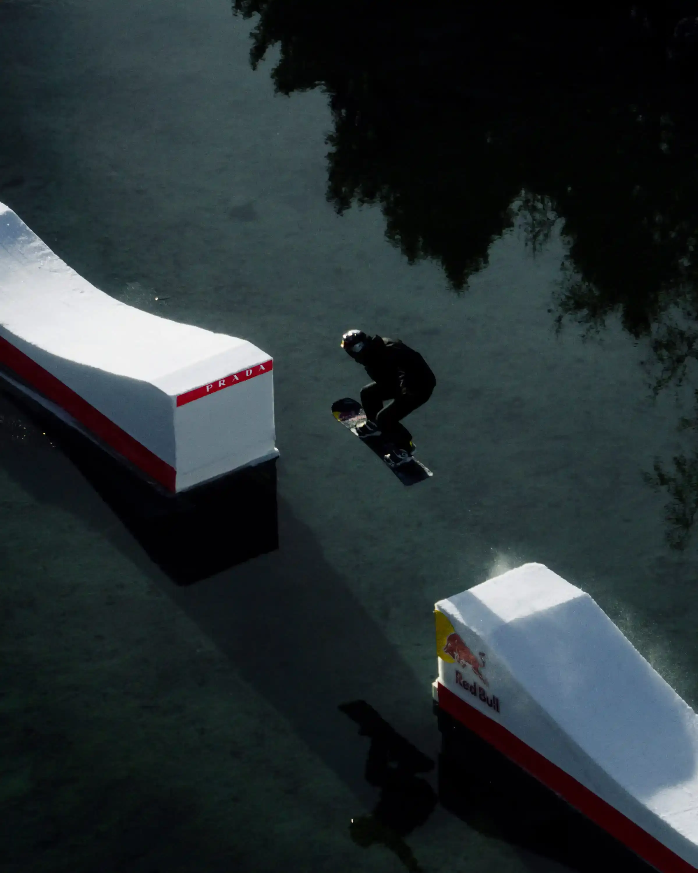 Prada Linea Rossa Snowboarder Pierre Vaultier riding on a narrow 50-centimeter track over a partially frozen lake in Serre-Chevalier. He wears black and red Prada Linea Rossa technical gear against a backdrop of snow-covered French Alps. (image 3 of 4)