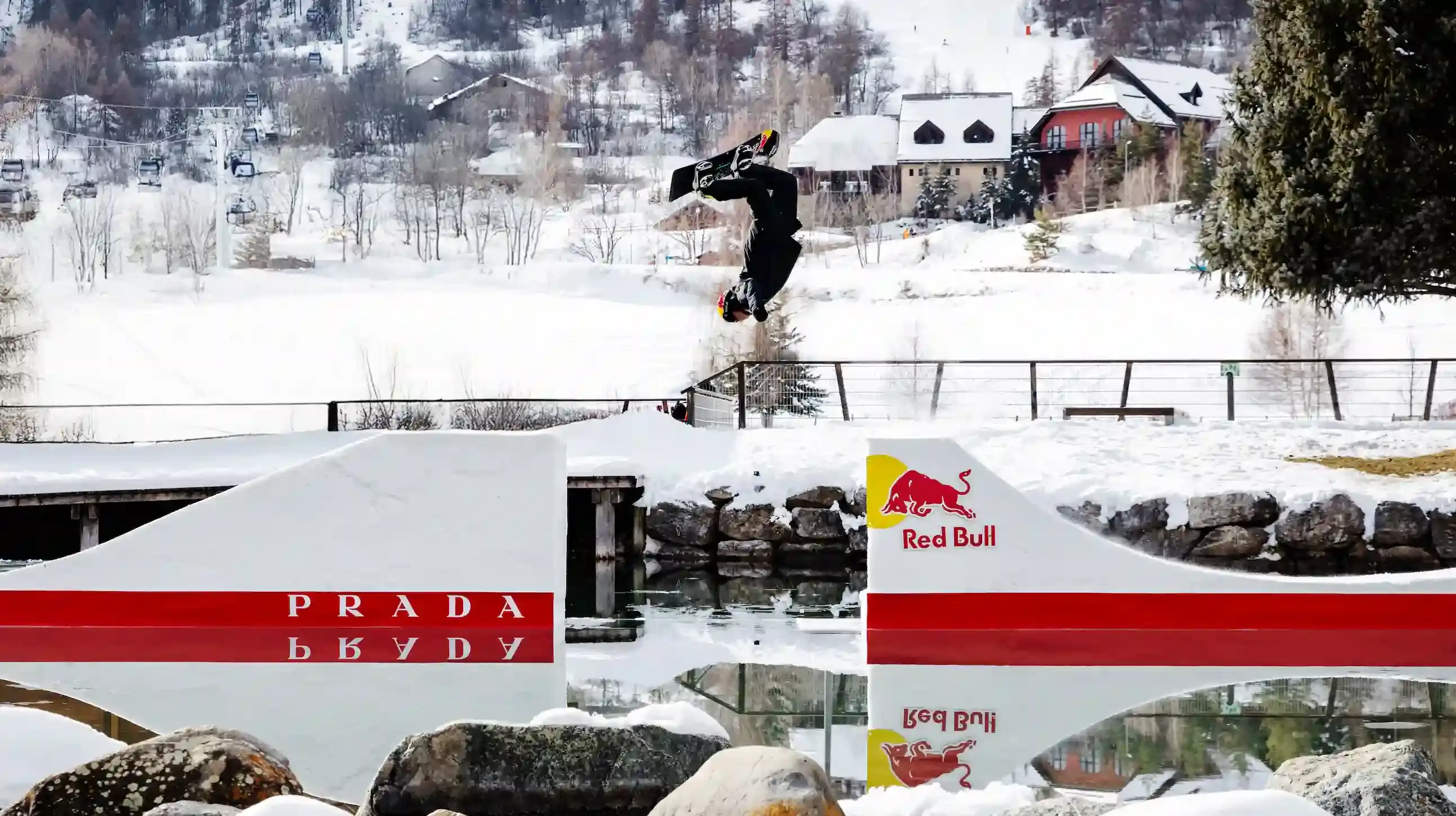Snowboarder Pierre Vaultier riding on a narrow 50-centimeter track over a partially frozen lake in Serre-Chevalier. He wears black and red Prada Linea Rossa technical gear against a backdrop of snow-covered French Alps. (image 2 of 2)