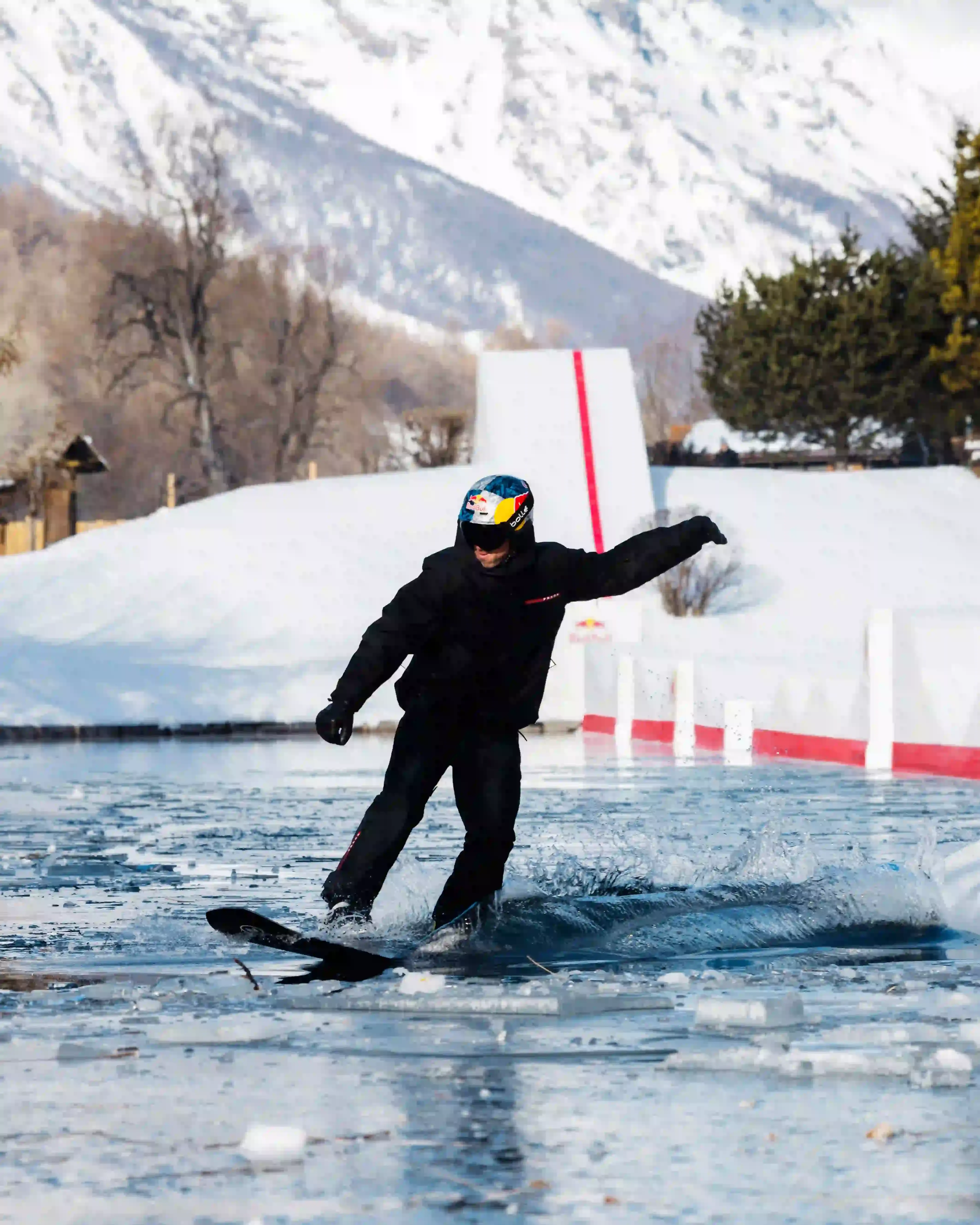 Prada Linea Rossa Snowboarder Pierre Vaultier riding on a narrow 50-centimeter track over a partially frozen lake in Serre-Chevalier. He wears black and red Prada Linea Rossa technical gear against a backdrop of snow-covered French Alps. (image 2 of 4)