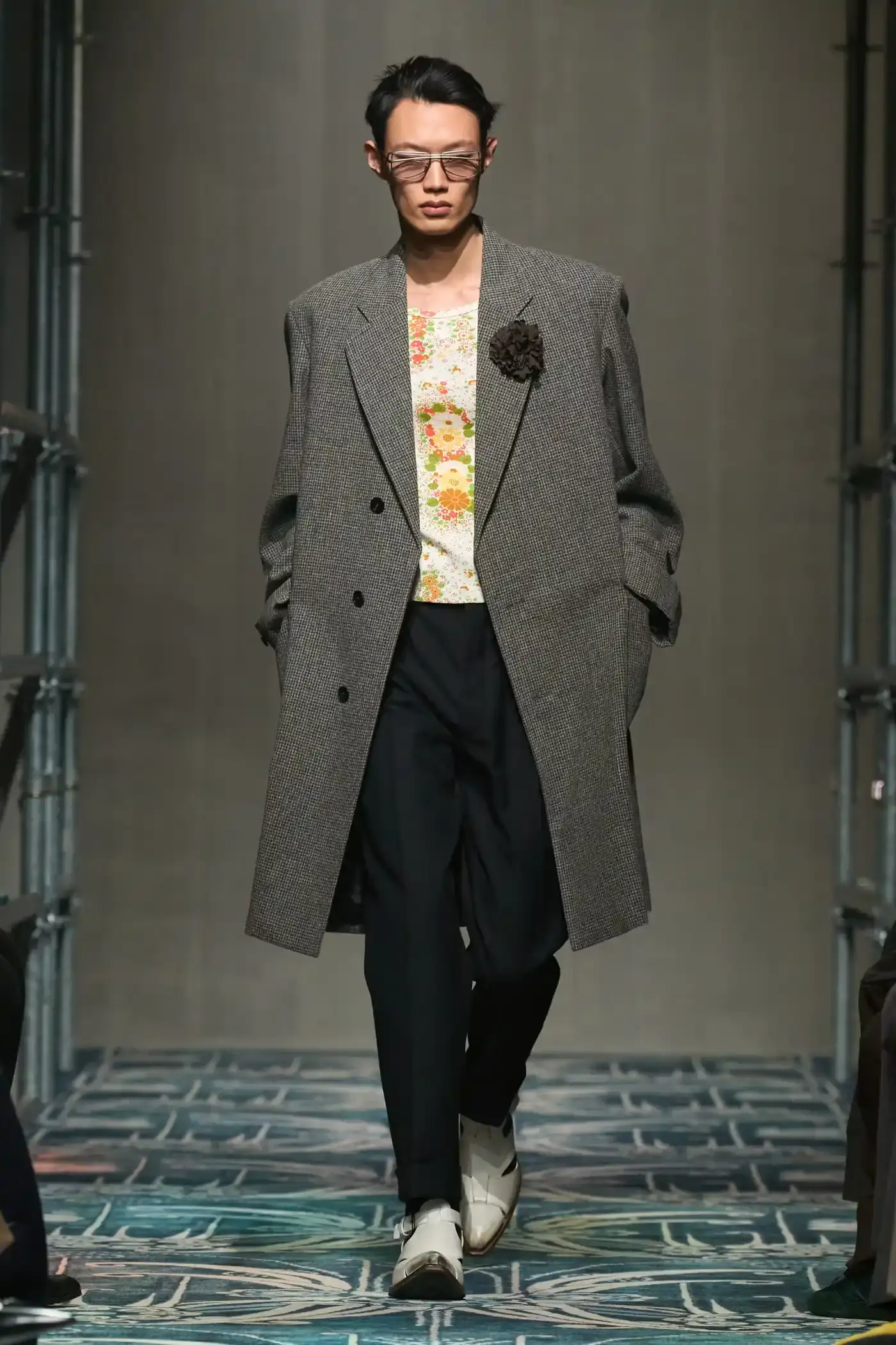 Filippo Fior/Gorunway.com/Vogue Runway A fashion show staged within a three-story industrial metal pipe structure at Fondazione Prada. The runway features male models walking through narrow corridors under shifting blue, red, and yellow lights. The industrial set is softened by a luxurious carpet. Models wear oversized coats, exposed chests with fur stoles, cottage-print tops, plaid trenches, and Western-influenced footwear. The collection demonstrates a mix of formal and casual elements, with unexpected combinations like floral brooches on tailored blazers. The space combines elements of both a nightclub and a traditional ballroom setting. (image 2 of 57)