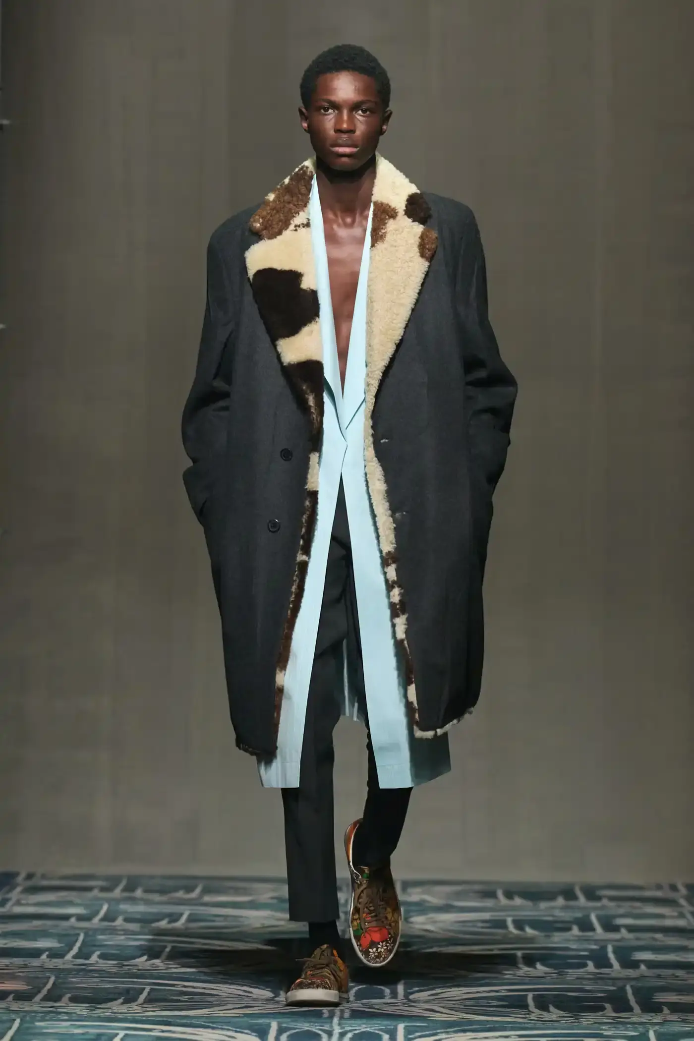 Filippo Fior/Gorunway.com/Vogue Runway A fashion show staged within a three-story industrial metal pipe structure at Fondazione Prada. The runway features male models walking through narrow corridors under shifting blue, red, and yellow lights. The industrial set is softened by a luxurious carpet. Models wear oversized coats, exposed chests with fur stoles, cottage-print tops, plaid trenches, and Western-influenced footwear. The collection demonstrates a mix of formal and casual elements, with unexpected combinations like floral brooches on tailored blazers. The space combines elements of both a nightclub and a traditional ballroom setting. (image 3 of 57)