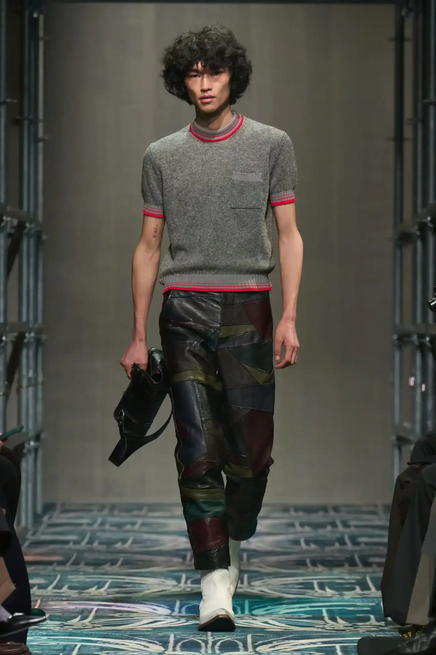 Filippo Fior/Gorunway.com/Vogue Runway A fashion show staged within a three-story industrial metal pipe structure at Fondazione Prada. The runway features male models walking through narrow corridors under shifting blue, red, and yellow lights. The industrial set is softened by a luxurious carpet. Models wear oversized coats, exposed chests with fur stoles, cottage-print tops, plaid trenches, and Western-influenced footwear. The collection demonstrates a mix of formal and casual elements, with unexpected combinations like floral brooches on tailored blazers. The space combines elements of both a nightclub and a traditional ballroom setting. (image 19 of 57)