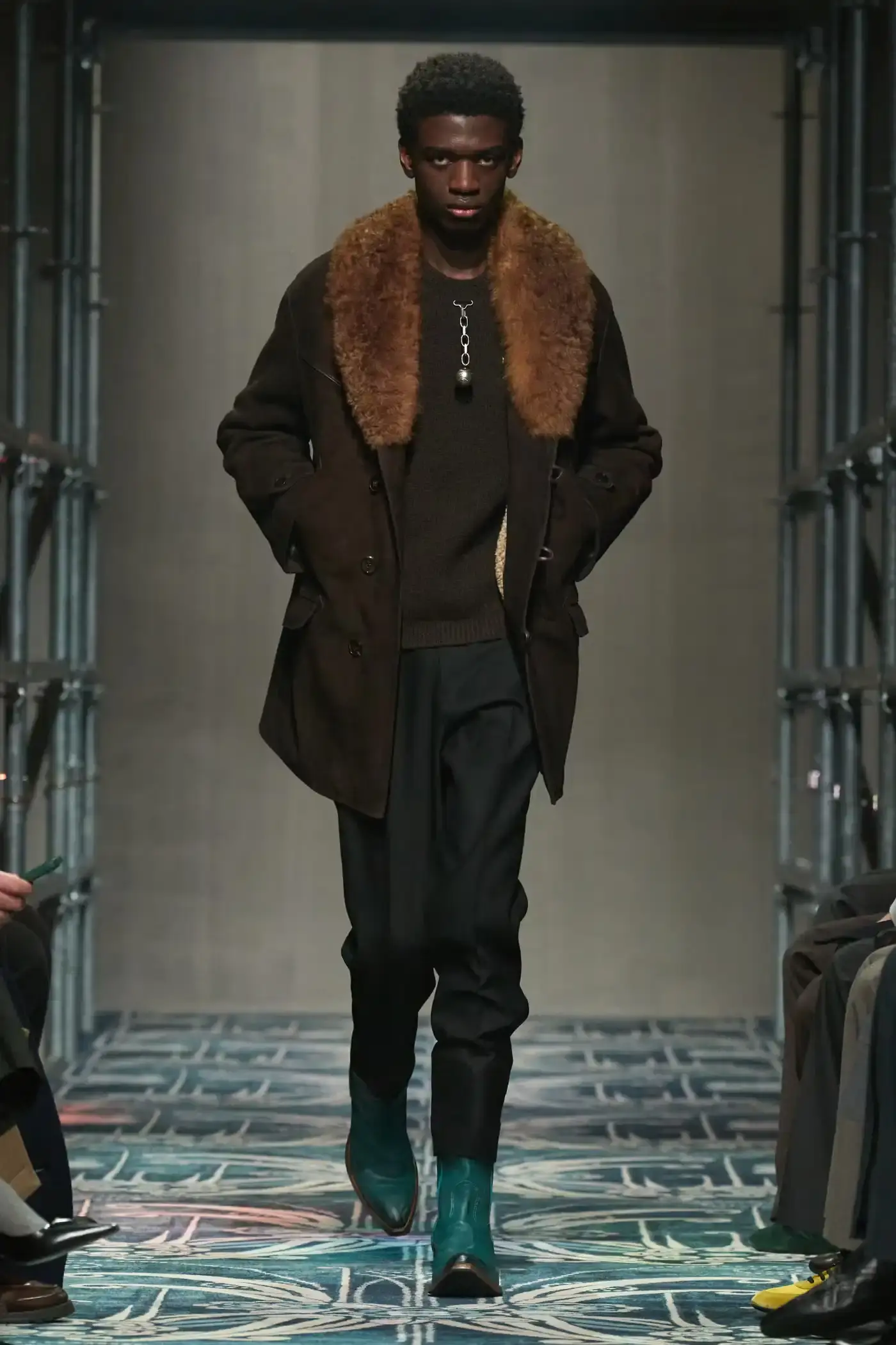 Filippo Fior/Gorunway.com/Vogue Runway A fashion show staged within a three-story industrial metal pipe structure at Fondazione Prada. The runway features male models walking through narrow corridors under shifting blue, red, and yellow lights. The industrial set is softened by a luxurious carpet. Models wear oversized coats, exposed chests with fur stoles, cottage-print tops, plaid trenches, and Western-influenced footwear. The collection demonstrates a mix of formal and casual elements, with unexpected combinations like floral brooches on tailored blazers. The space combines elements of both a nightclub and a traditional ballroom setting. (image 14 of 57)