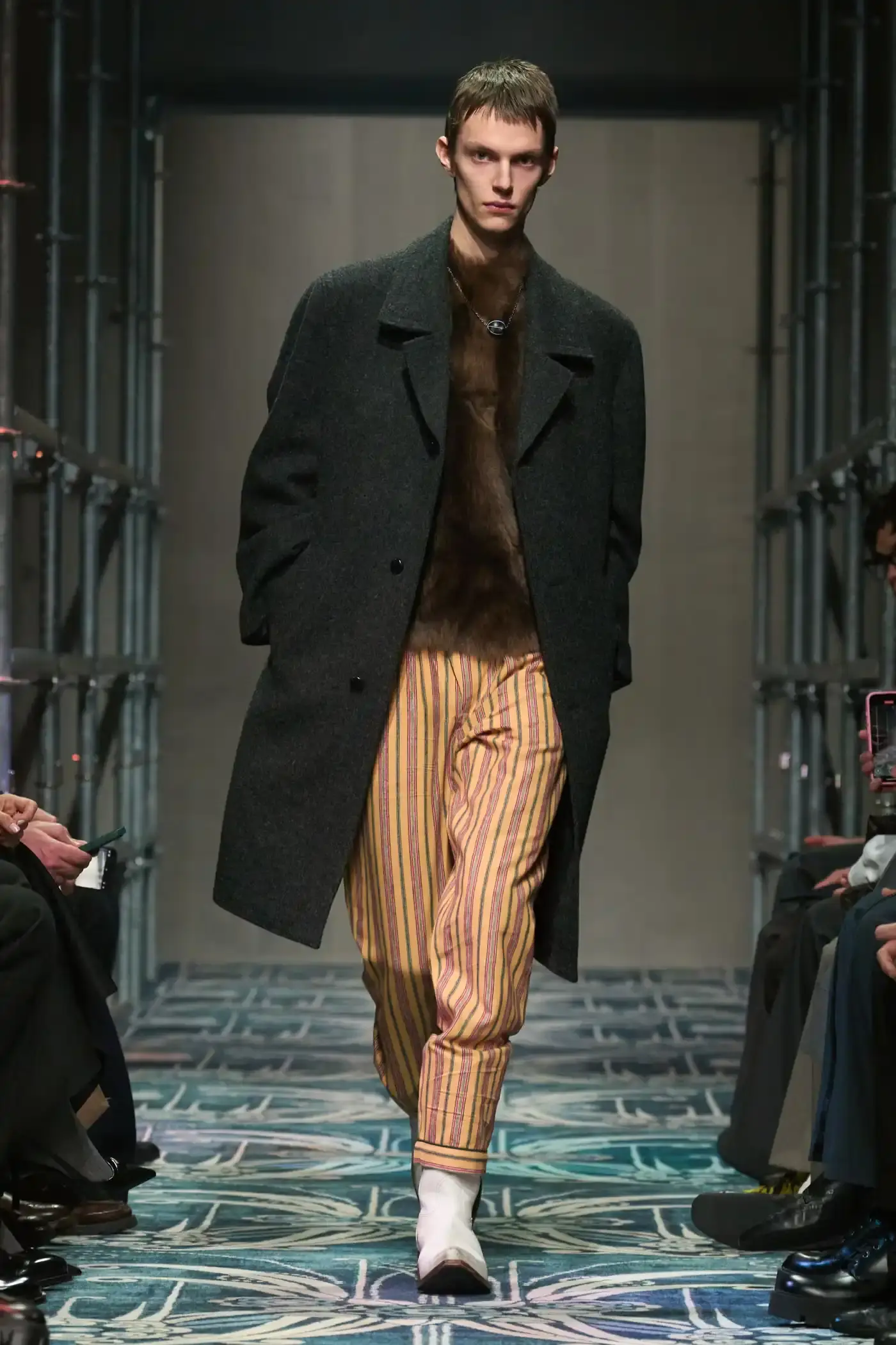 Filippo Fior/Gorunway.com/Vogue Runway A fashion show staged within a three-story industrial metal pipe structure at Fondazione Prada. The runway features male models walking through narrow corridors under shifting blue, red, and yellow lights. The industrial set is softened by a luxurious carpet. Models wear oversized coats, exposed chests with fur stoles, cottage-print tops, plaid trenches, and Western-influenced footwear. The collection demonstrates a mix of formal and casual elements, with unexpected combinations like floral brooches on tailored blazers. The space combines elements of both a nightclub and a traditional ballroom setting. (image 26 of 57)
