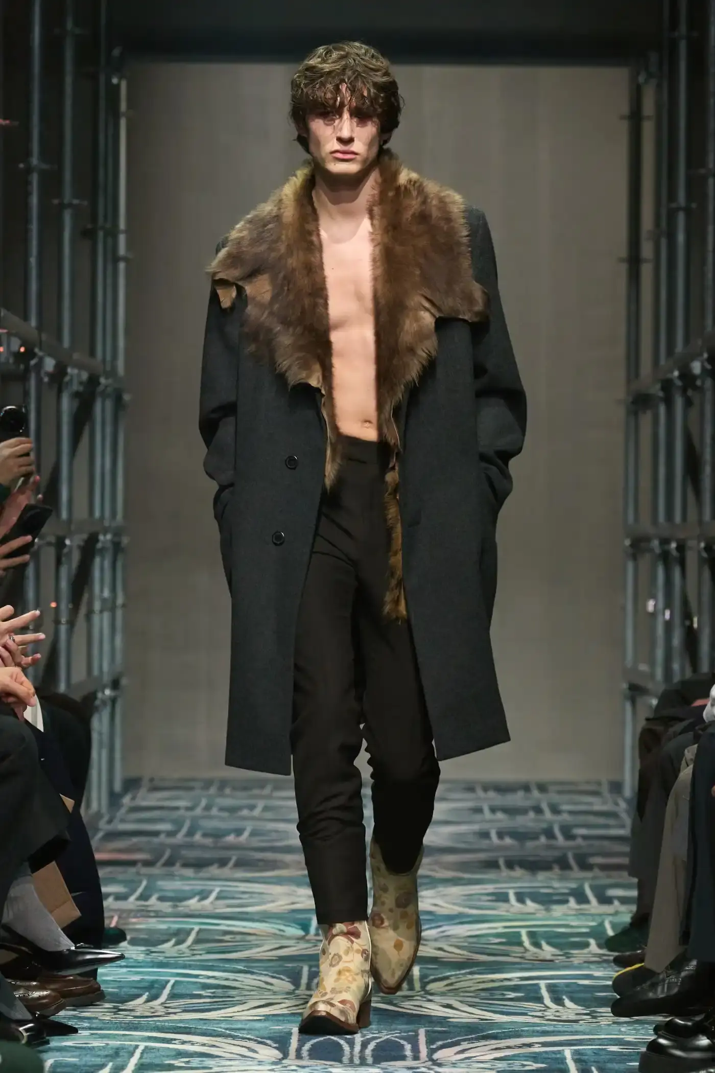Filippo Fior/Gorunway.com/Vogue Runway A fashion show staged within a three-story industrial metal pipe structure at Fondazione Prada. The runway features male models walking through narrow corridors under shifting blue, red, and yellow lights. The industrial set is softened by a luxurious carpet. Models wear oversized coats, exposed chests with fur stoles, cottage-print tops, plaid trenches, and Western-influenced footwear. The collection demonstrates a mix of formal and casual elements, with unexpected combinations like floral brooches on tailored blazers. The space combines elements of both a nightclub and a traditional ballroom setting.