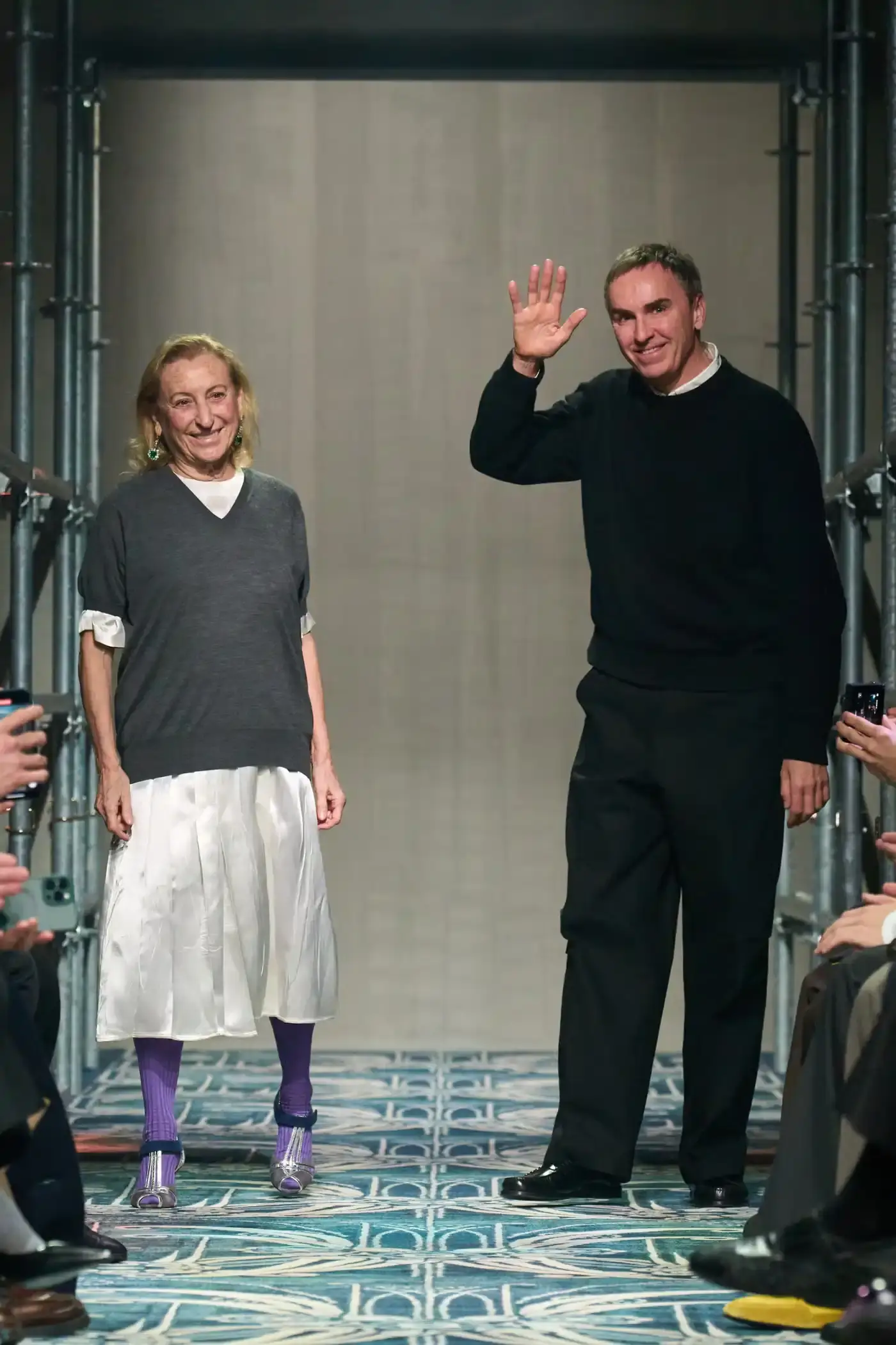 Filippo Fior/Gorunway.com/Vogue Runway A fashion show staged within a three-story industrial metal pipe structure at Fondazione Prada. The runway features male models walking through narrow corridors under shifting blue, red, and yellow lights. The industrial set is softened by a luxurious carpet. Models wear oversized coats, exposed chests with fur stoles, cottage-print tops, plaid trenches, and Western-influenced footwear. The collection demonstrates a mix of formal and casual elements, with unexpected combinations like floral brooches on tailored blazers. The space combines elements of both a nightclub and a traditional ballroom setting. (image 57 of 57)