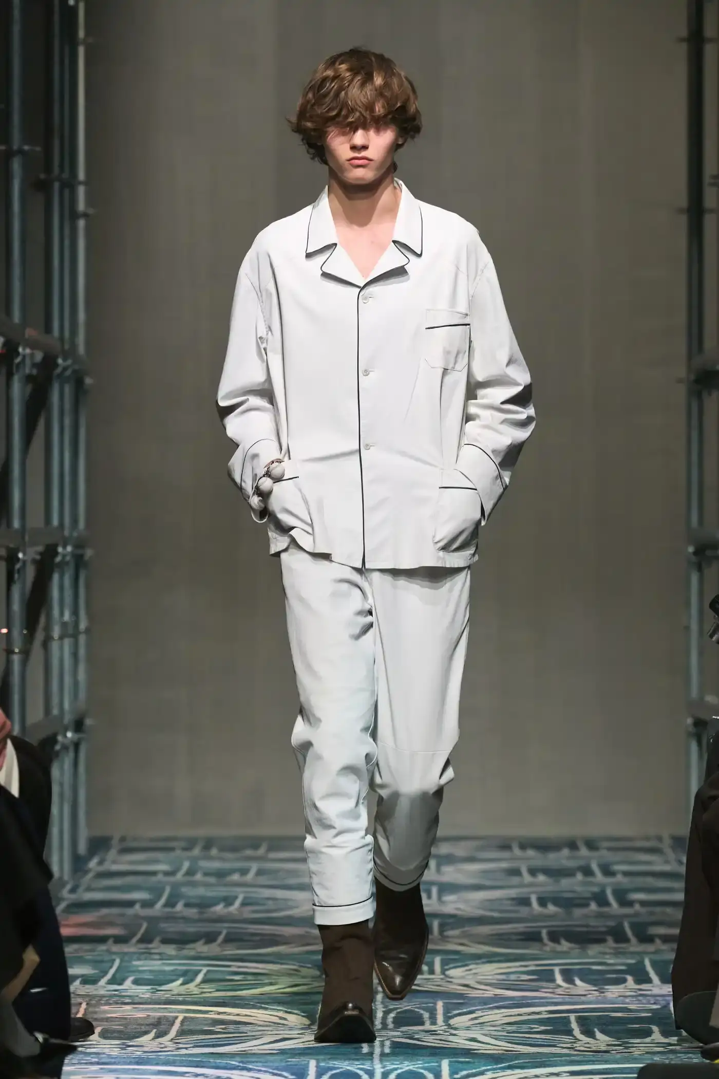 Filippo Fior/Gorunway.com/Vogue Runway A fashion show staged within a three-story industrial metal pipe structure at Fondazione Prada. The runway features male models walking through narrow corridors under shifting blue, red, and yellow lights. The industrial set is softened by a luxurious carpet. Models wear oversized coats, exposed chests with fur stoles, cottage-print tops, plaid trenches, and Western-influenced footwear. The collection demonstrates a mix of formal and casual elements, with unexpected combinations like floral brooches on tailored blazers. The space combines elements of both a nightclub and a traditional ballroom setting. (image 42 of 57)