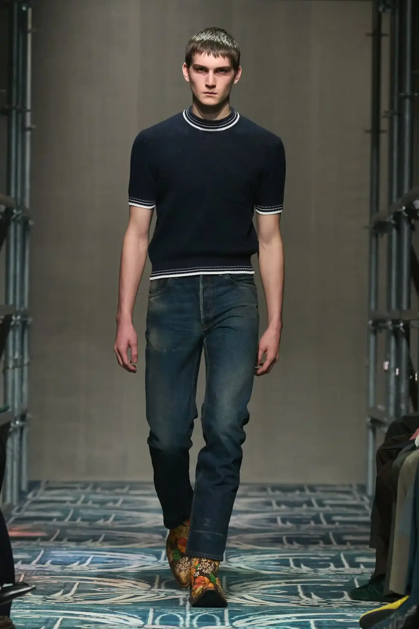 Filippo Fior/Gorunway.com/Vogue Runway A fashion show staged within a three-story industrial metal pipe structure at Fondazione Prada. The runway features male models walking through narrow corridors under shifting blue, red, and yellow lights. The industrial set is softened by a luxurious carpet. Models wear oversized coats, exposed chests with fur stoles, cottage-print tops, plaid trenches, and Western-influenced footwear. The collection demonstrates a mix of formal and casual elements, with unexpected combinations like floral brooches on tailored blazers. The space combines elements of both a nightclub and a traditional ballroom setting. (image 4 of 57)