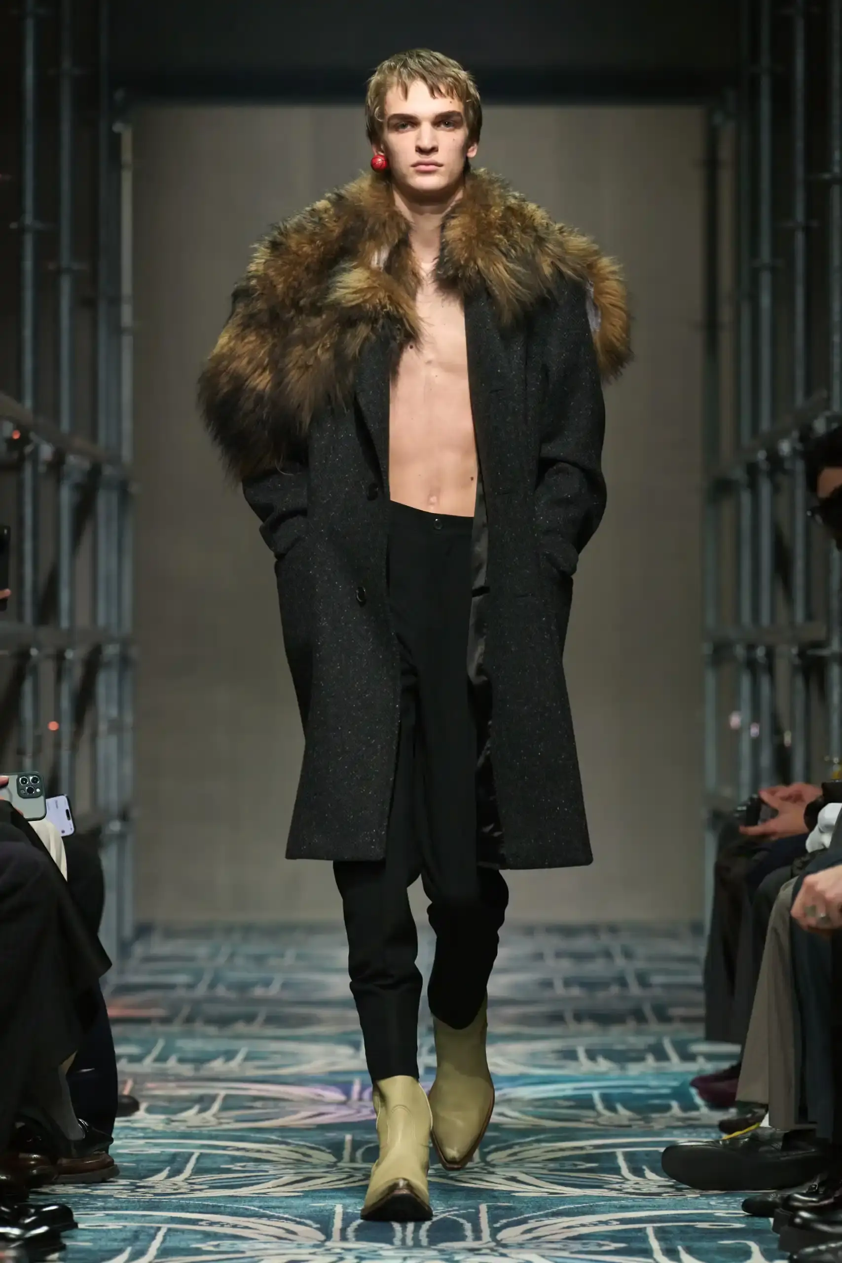 Filippo Fior/Gorunway.com/Vogue Runway A fashion show staged within a three-story industrial metal pipe structure at Fondazione Prada. The runway features male models walking through narrow corridors under shifting blue, red, and yellow lights. The industrial set is softened by a luxurious carpet. Models wear oversized coats, exposed chests with fur stoles, cottage-print tops, plaid trenches, and Western-influenced footwear. The collection demonstrates a mix of formal and casual elements, with unexpected combinations like floral brooches on tailored blazers. The space combines elements of both a nightclub and a traditional ballroom setting. (image 55 of 57)