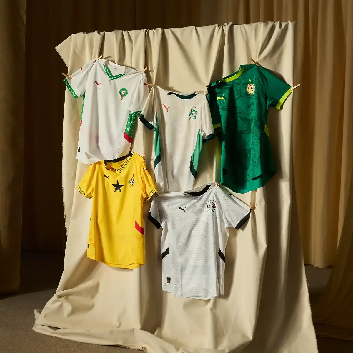 Puma PUMA's 2025 AFCON collection displayed on mannequins, showing home and away jerseys for Morocco and Ghana national teams. The Morocco kit features geometric patterns in red and white, while Ghana's uniforms combine traditional symbols with yellow and white colorways.