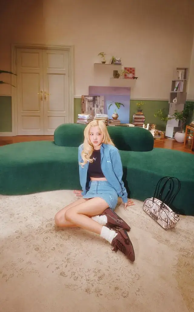ROSE FRONTS PUMA SPEEDCAT CROC CAMPAIGN A blonde woman wearing blue denim sits on a modern chair in a retro room. She wears brown PUMA Speedcat Croc shoes next to a matching bag.