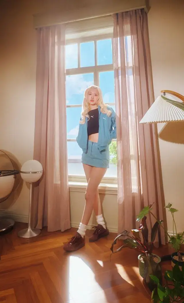 ROSE FRONTS PUMA SPEEDCAT CROC CAMPAIGN A blonde woman wearing blue denim sits on a modern chair in a retro room. She wears brown PUMA Speedcat Croc shoes next to a matching bag. (image 2 of 4)