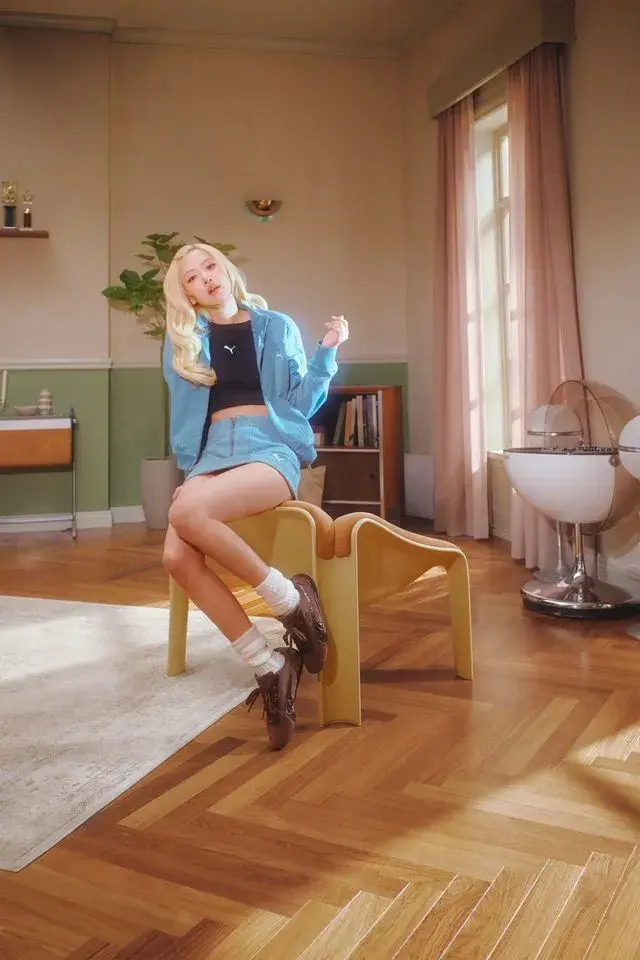 ROSE FRONTS PUMA SPEEDCAT CROC CAMPAIGN A blonde woman wearing blue denim sits on a modern chair in a retro room. She wears brown PUMA Speedcat Croc shoes next to a matching bag. (image 3 of 4)