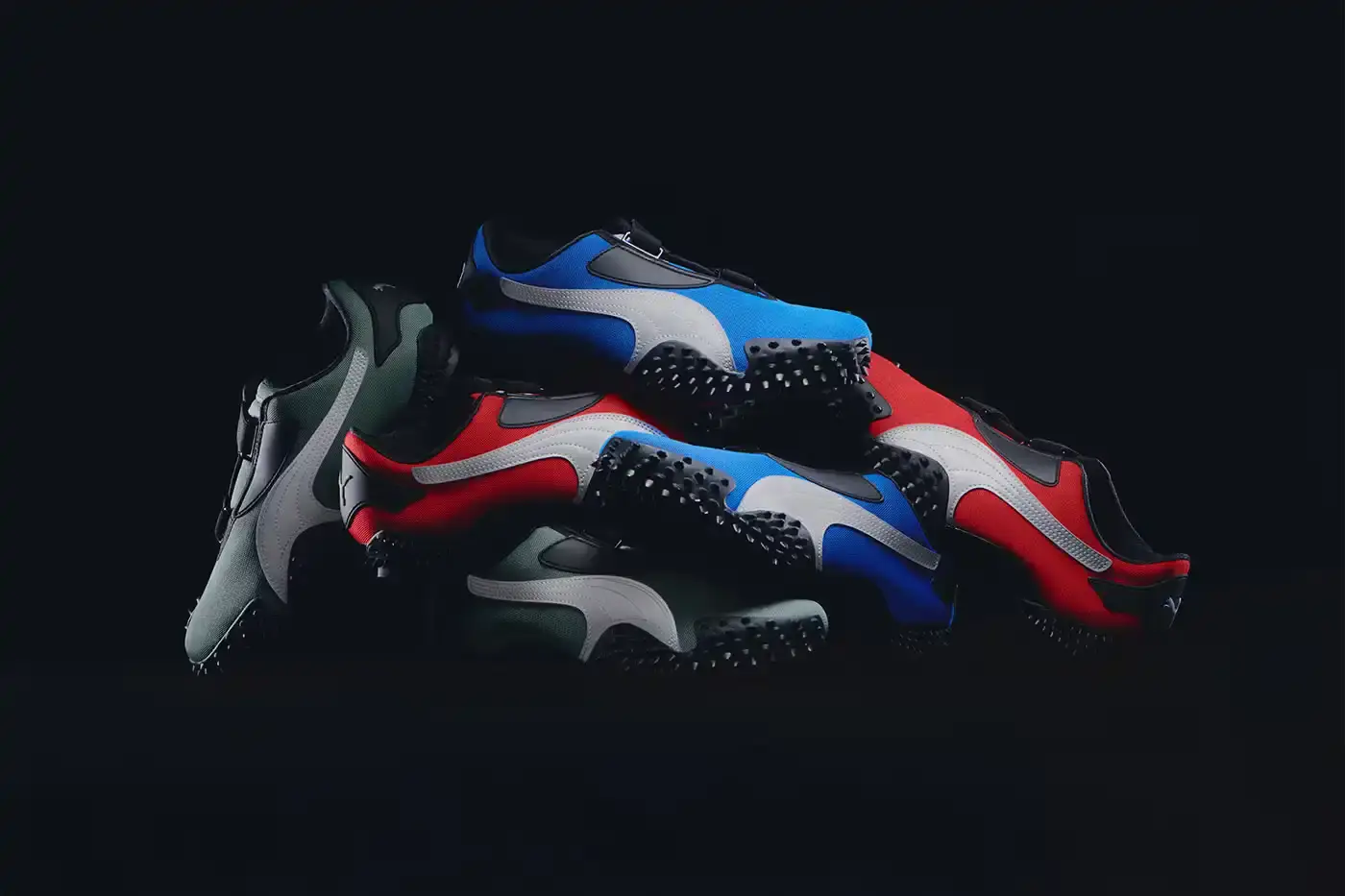 Puma PUMA Mostro OG sneakers displayed in three colorways - Team Royal blue with silver accents, Green Moon green with silver detailing, and All Time Red with silver trim. Each shoe features the model's distinctive spiked sole, mesh upper, and hook-and-loop fastening system.