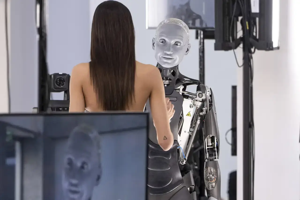 A fashion runway scene showing model Yasmin Wijnaldum in a sculptural jeweled corset standing next to Ameca, a humanoid robot with distinctive mechanical features, both wearing Giuseppe di Morabito designs against a dark backdrop that highlights the contrast between human and machine.