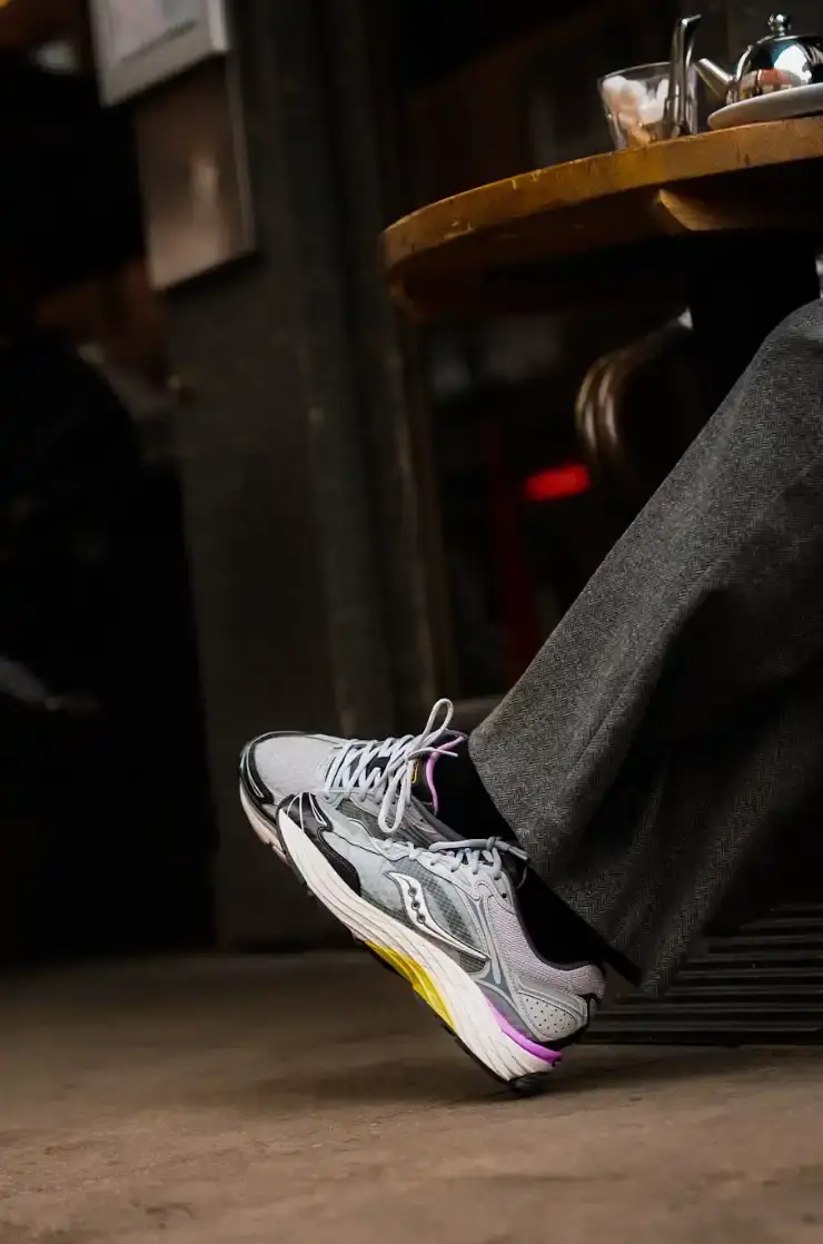 SAUCONY PROGRID OMNI 9 SS26 RELEASE An image showing a person sitting on a city street wearing Saucony ProGrid Omni 9 sneakers in a dark colorway with pink accents. (image 6 of 6)