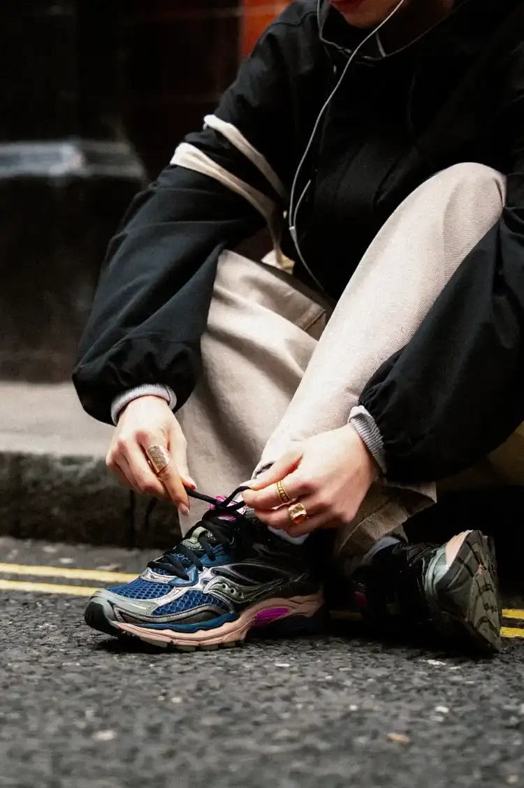 SAUCONY PROGRID OMNI 9 SS26 RELEASE An image showing a person sitting on a city street wearing Saucony ProGrid Omni 9 sneakers in a dark colorway with pink accents. (image 2 of 6)