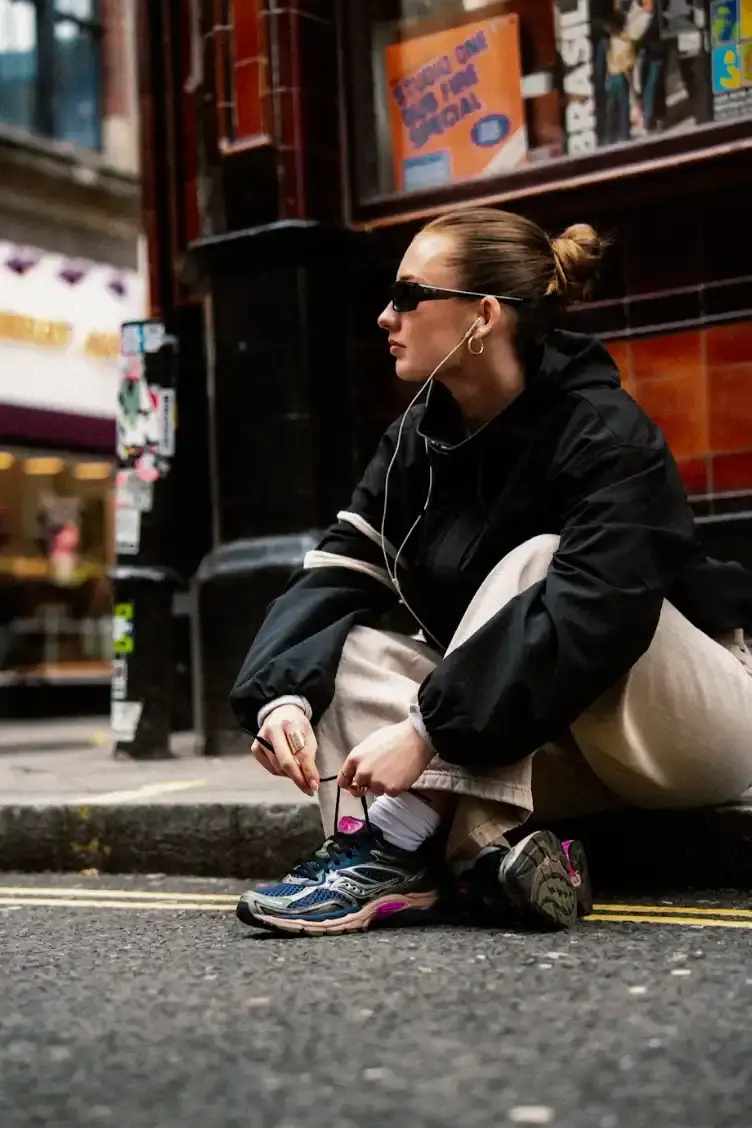 SAUCONY PROGRID OMNI 9 SS26 RELEASE An image showing a person sitting on a city street wearing Saucony ProGrid Omni 9 sneakers in a dark colorway with pink accents.