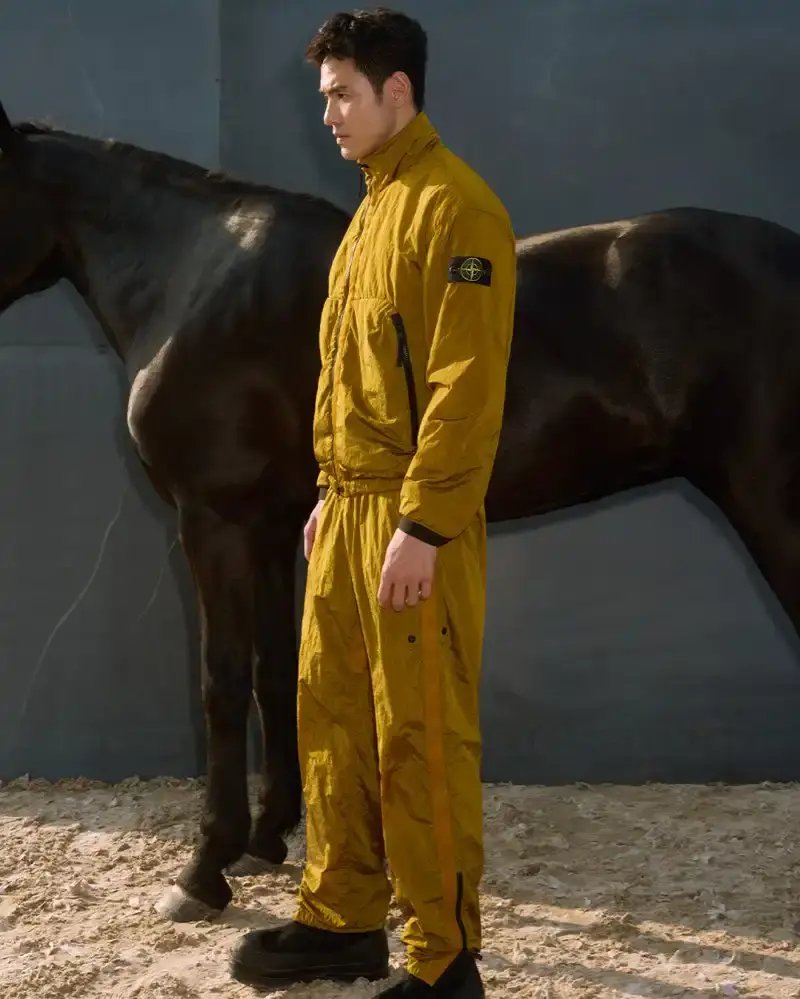 STONE ISLAND LUNAR NEW YEAR HORSE COLLECTION Stone Island introduces a Lunar New Year collection for the Year of the Horse, featuring a stand collar jacket, joggers, cap, and sling bag in NYLON METAL fabric with gold and dark brown colors.