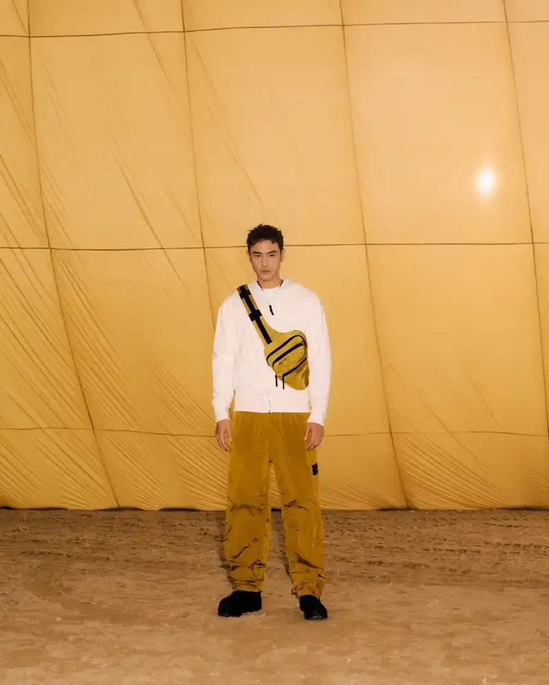 STONE ISLAND LUNAR NEW YEAR HORSE COLLECTION Stone Island introduces a Lunar New Year collection for the Year of the Horse, featuring a stand collar jacket, joggers, cap, and sling bag in NYLON METAL fabric with gold and dark brown colors. (image 5 of 10)