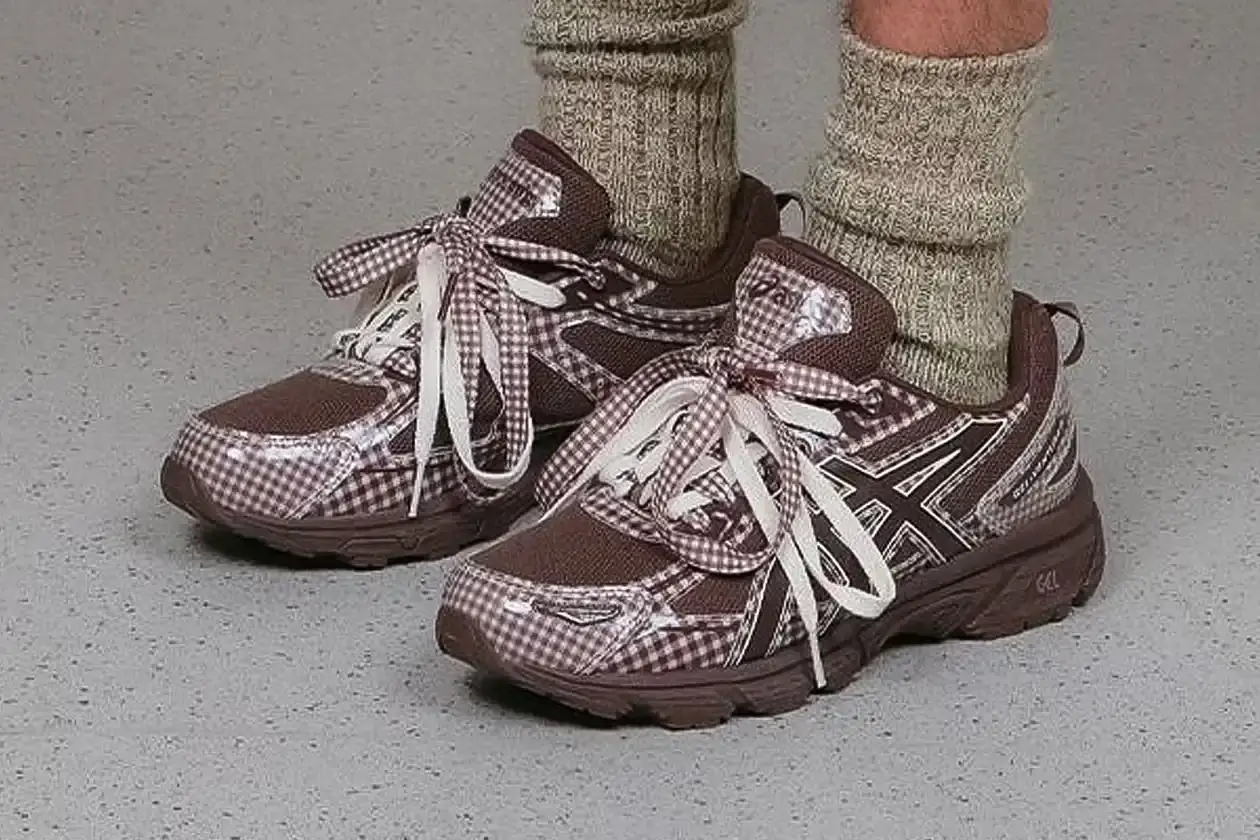 ASICS GEL-VENTURE 6 GETS GINGHAM MAKEOVER FROM STORY MFG. photo of the collaboration between sustainable fashion brand STORY mfg. and ASICS on the GEL-VENTURE 6 sneaker. It details the two gingham colorways ("Reddish Brown" and "Vanilla Violet Quartz"), the unique woven lacing system inspired by Baghdad skaters, and pricing ($133 USD). The article also describes the two-day launch event at The Truman Brewery in London on April 18-19, where visitors can contribute embroidery to a community art project while enjoying refreshments. The article provides purchase information and explains STORY mfg.'s sustainable fashion background.