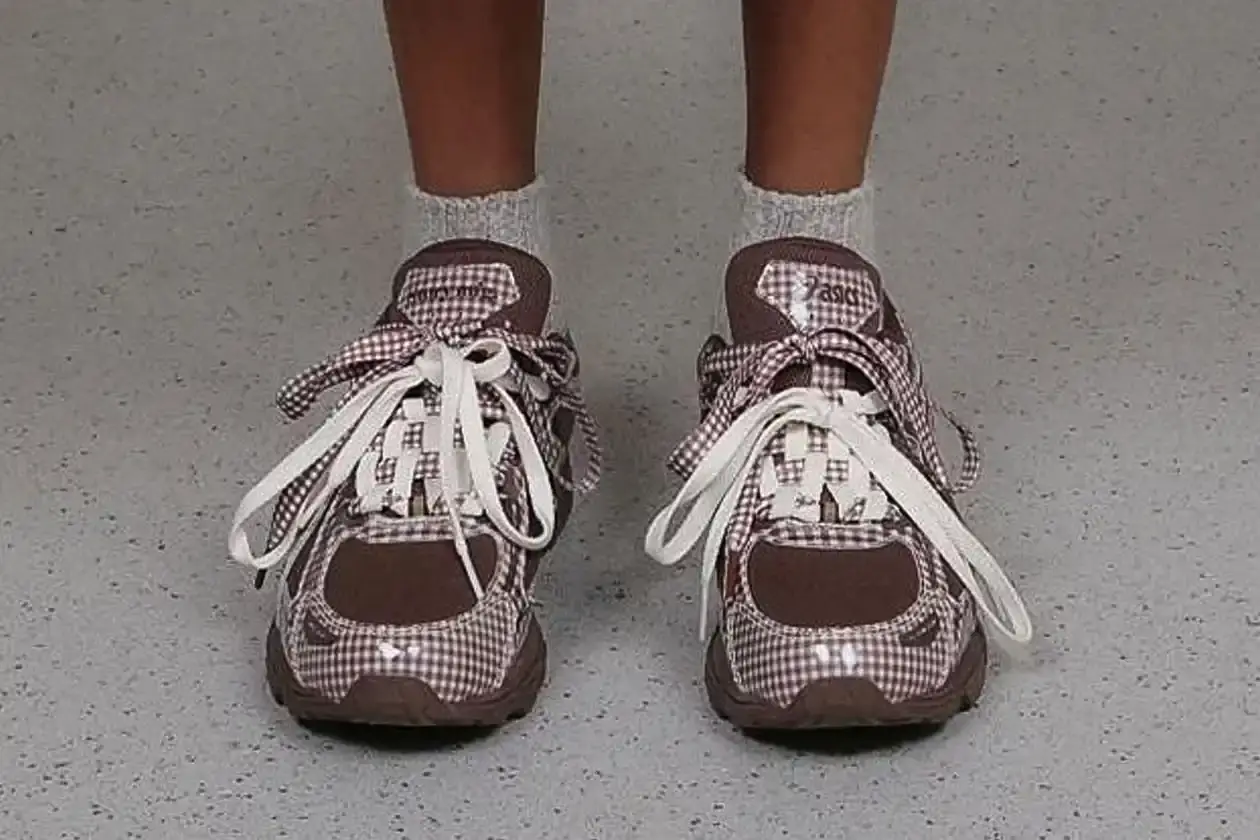 ASICS GEL-VENTURE 6 GETS GINGHAM MAKEOVER FROM STORY MFG. photo of the collaboration between sustainable fashion brand STORY mfg. and ASICS on the GEL-VENTURE 6 sneaker. It details the two gingham colorways ("Reddish Brown" and "Vanilla Violet Quartz"), the unique woven lacing system inspired by Baghdad skaters, and pricing ($133 USD). The article also describes the two-day launch event at The Truman Brewery in London on April 18-19, where visitors can contribute embroidery to a community art project while enjoying refreshments. The article provides purchase information and explains STORY mfg.'s sustainable fashion background. (image 3 of 14)