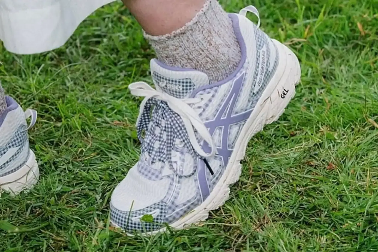 ASICS GEL-VENTURE 6 GETS GINGHAM MAKEOVER FROM STORY MFG. photo of the collaboration between sustainable fashion brand STORY mfg. and ASICS on the GEL-VENTURE 6 sneaker. It details the two gingham colorways ("Reddish Brown" and "Vanilla Violet Quartz"), the unique woven lacing system inspired by Baghdad skaters, and pricing ($133 USD). The article also describes the two-day launch event at The Truman Brewery in London on April 18-19, where visitors can contribute embroidery to a community art project while enjoying refreshments. The article provides purchase information and explains STORY mfg.'s sustainable fashion background. (image 14 of 14)