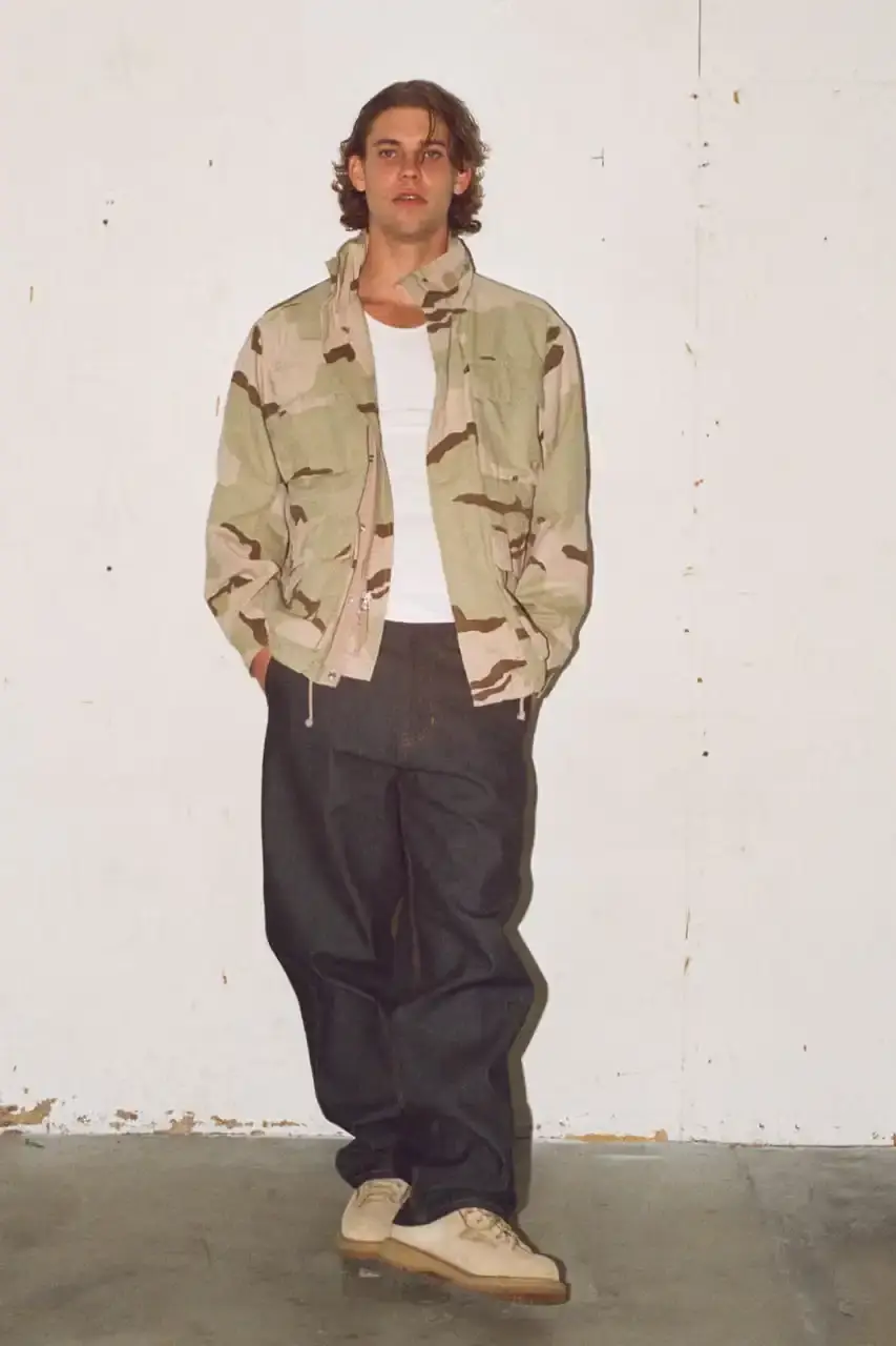 Antosh Cimoszko Stüssy Spring 2025 collection lookbook featuring an urban model wearing an oversized camouflage jacket, baggy denim trousers, and wide-cut shorts in muted green and blue tones. (image 7 of 26)