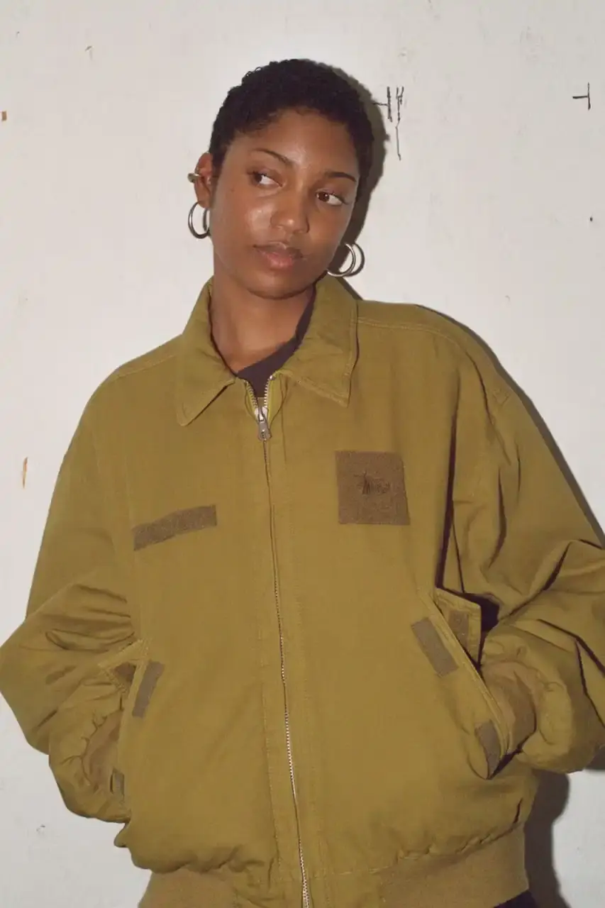 Antosh Cimoszko Stüssy Spring 2025 collection lookbook featuring an urban model wearing an oversized camouflage jacket, baggy denim trousers, and wide-cut shorts in muted green and blue tones. (image 9 of 26)