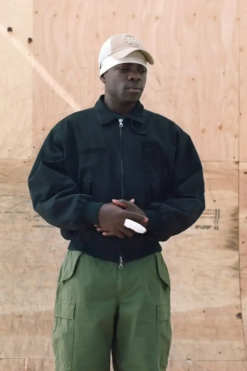 Antosh Cimoszko Stüssy Spring 2025 collection lookbook featuring an urban model wearing an oversized camouflage jacket, baggy denim trousers, and wide-cut shorts in muted green and blue tones. (image 18 of 26)