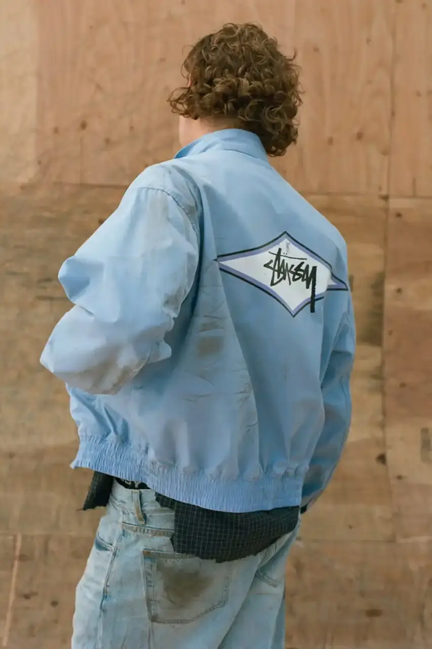 Antosh Cimoszko Stüssy Spring 2025 collection lookbook featuring an urban model wearing an oversized camouflage jacket, baggy denim trousers, and wide-cut shorts in muted green and blue tones. (image 4 of 26)
