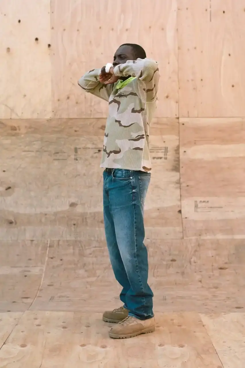 Antosh Cimoszko Stüssy Spring 2025 collection lookbook featuring an urban model wearing an oversized camouflage jacket, baggy denim trousers, and wide-cut shorts in muted green and blue tones. (image 22 of 26)