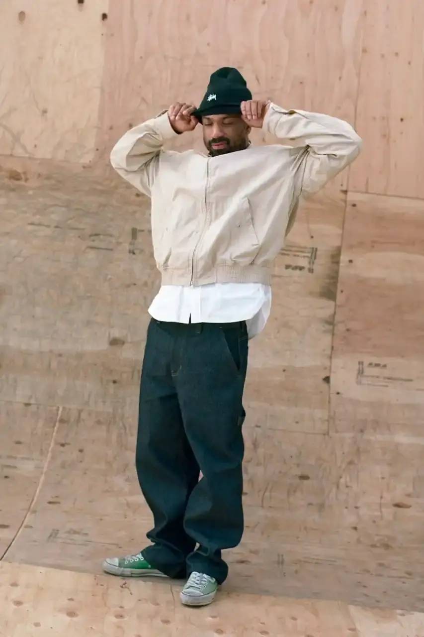 Antosh Cimoszko Stüssy Spring 2025 collection lookbook featuring an urban model wearing an oversized camouflage jacket, baggy denim trousers, and wide-cut shorts in muted green and blue tones. (image 17 of 26)