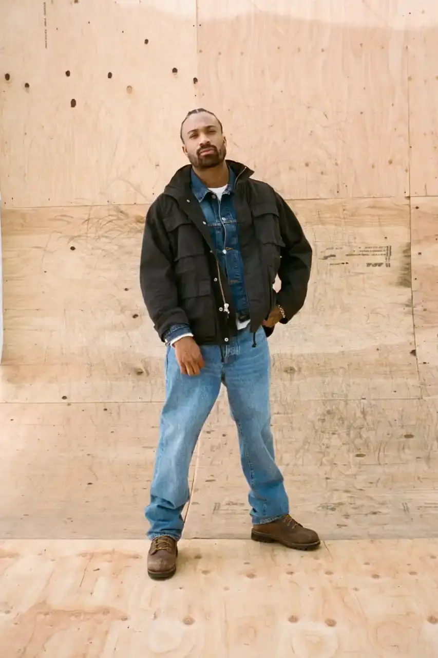 Antosh Cimoszko Stüssy Spring 2025 collection lookbook featuring an urban model wearing an oversized camouflage jacket, baggy denim trousers, and wide-cut shorts in muted green and blue tones. (image 19 of 26)
