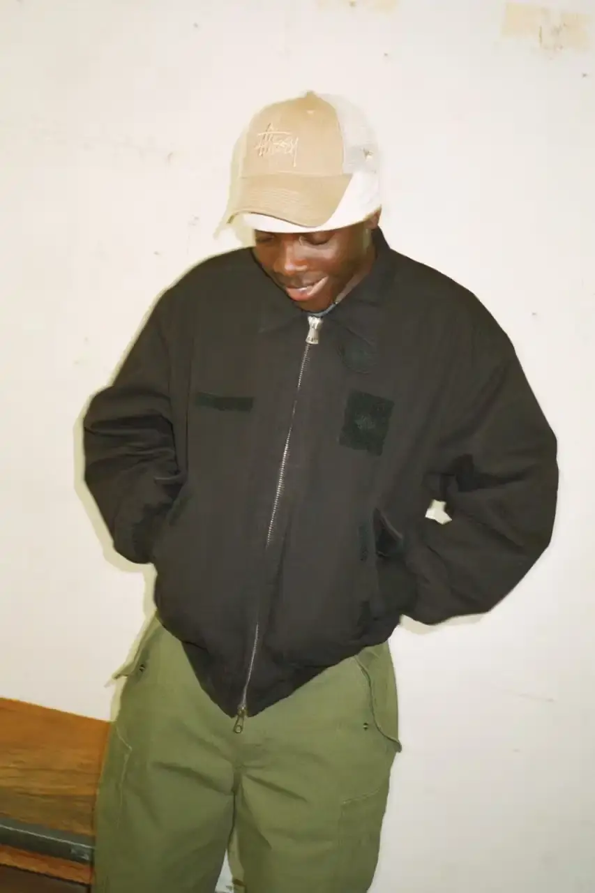 Antosh Cimoszko Stüssy Spring 2025 collection lookbook featuring an urban model wearing an oversized camouflage jacket, baggy denim trousers, and wide-cut shorts in muted green and blue tones. (image 11 of 26)